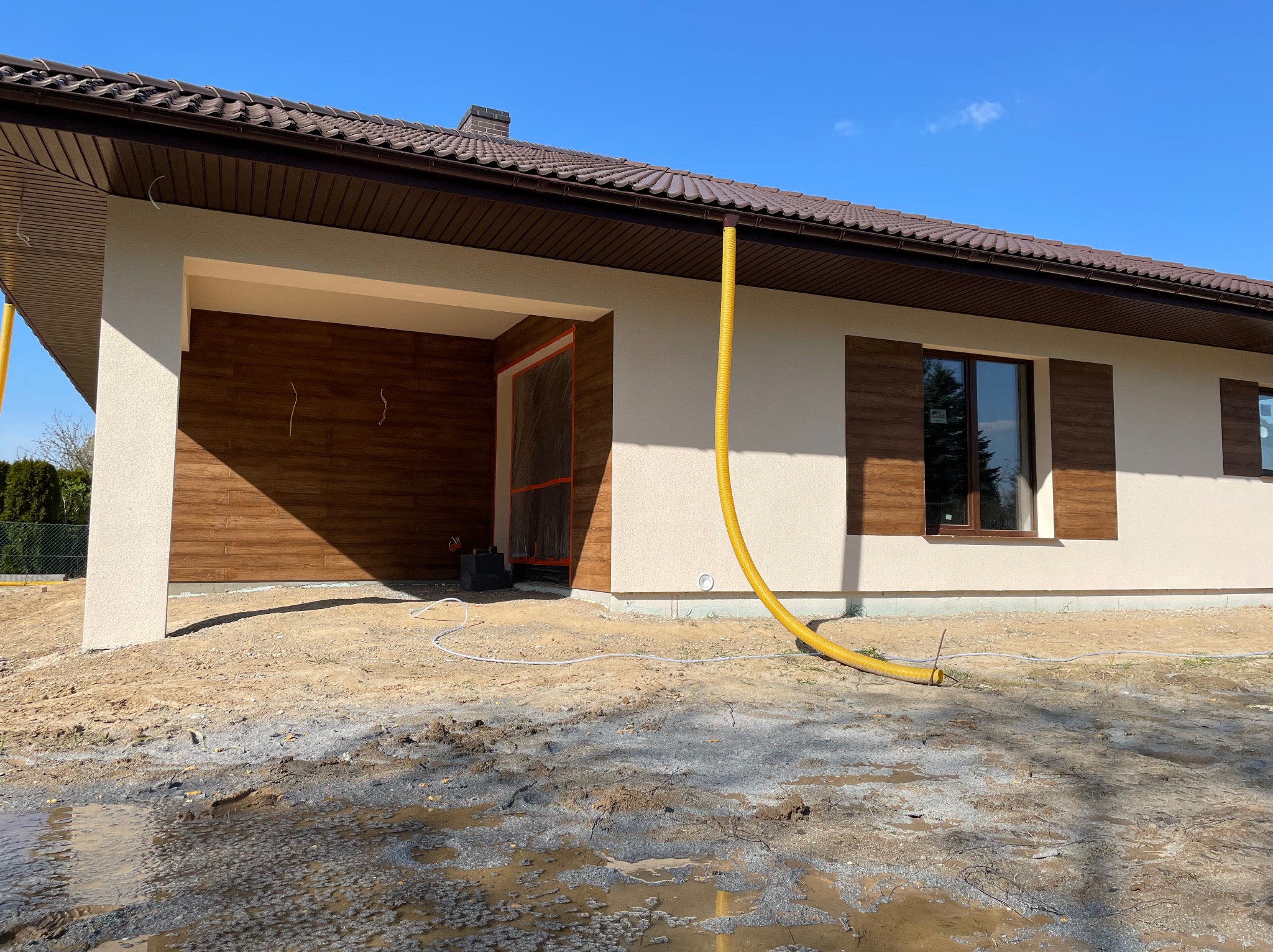 Dom jednorodzinny w trakcie prac elewacyjnych, widoczna żółta rura wentylacyjna, świeżo otynkowane ściany w jasnym odcieniu, brązowe okiennice i fragment drewnianej elewacji garażu, teren wokół...