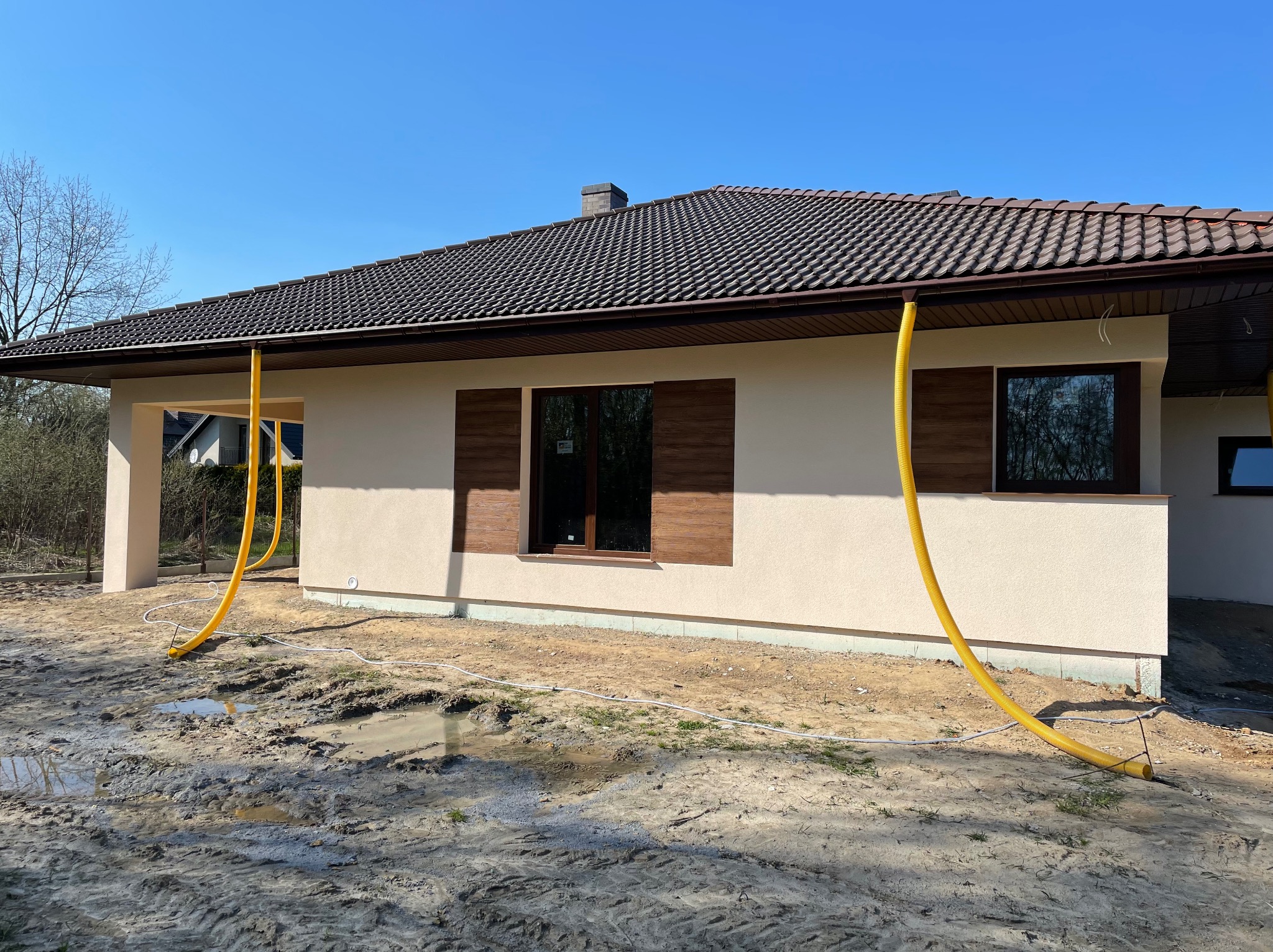 Nowo otynkowany dom jednorodzinny z brązową dachówką i oknami z imitacją drewna, widoczne żółte rury wentylacyjne przy elewacji, teren wokół domu częściowo błotnisty.