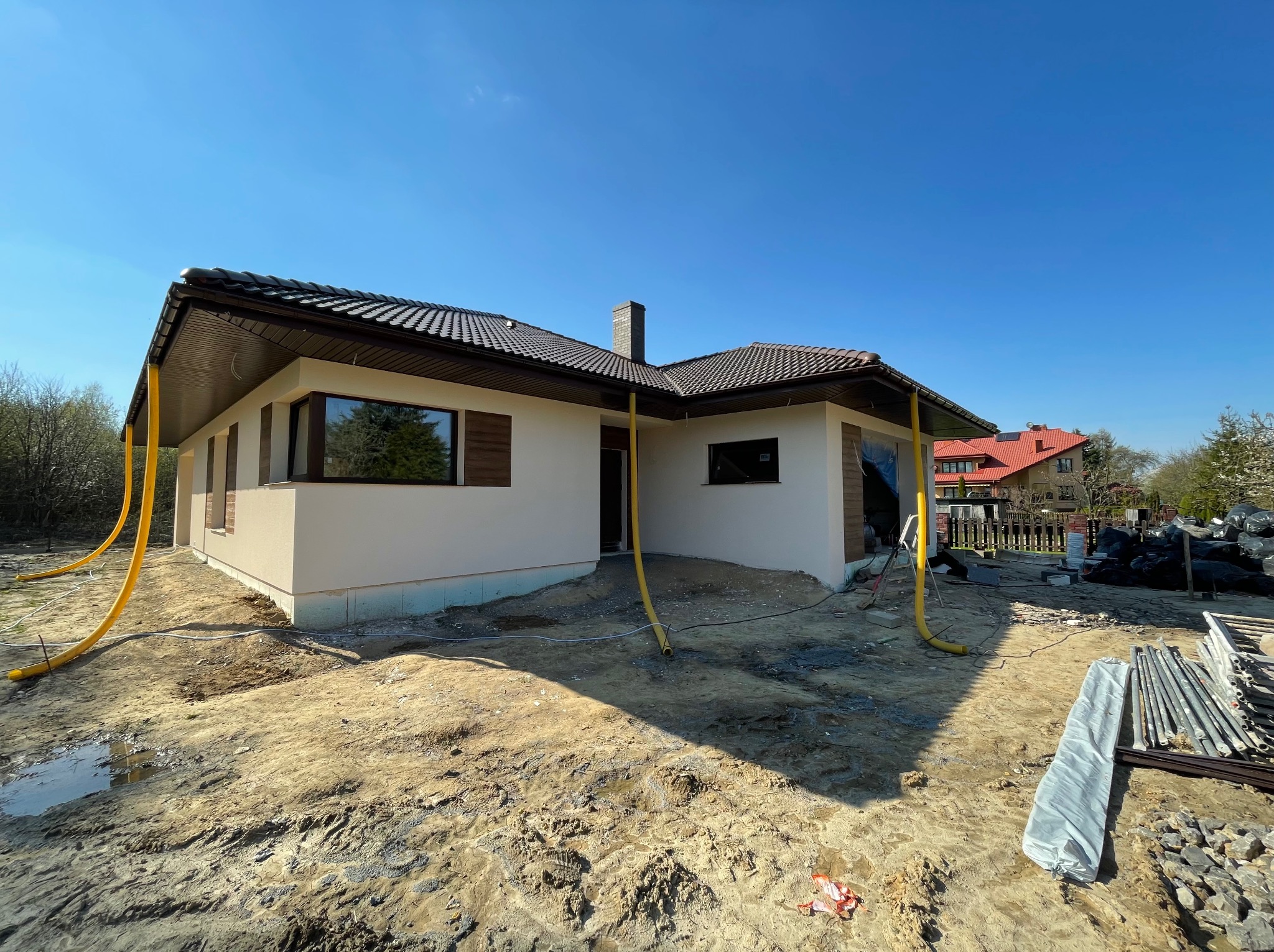 Niski, parterowy dom z jasną elewacją, brązowym dachem i oknami z drewnianymi okiennicami, widoczny w trakcie prac wykończeniowych, z żółtymi rurami wentylacyjnymi na zewnątrz i rozkopanym terenem...
