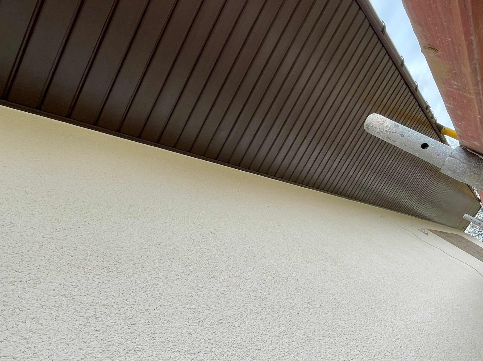 Fragment elewacji z beżowym tynkiem strukturalnym i brązowym podbitką dachową, widoczne rusztowanie z boku.