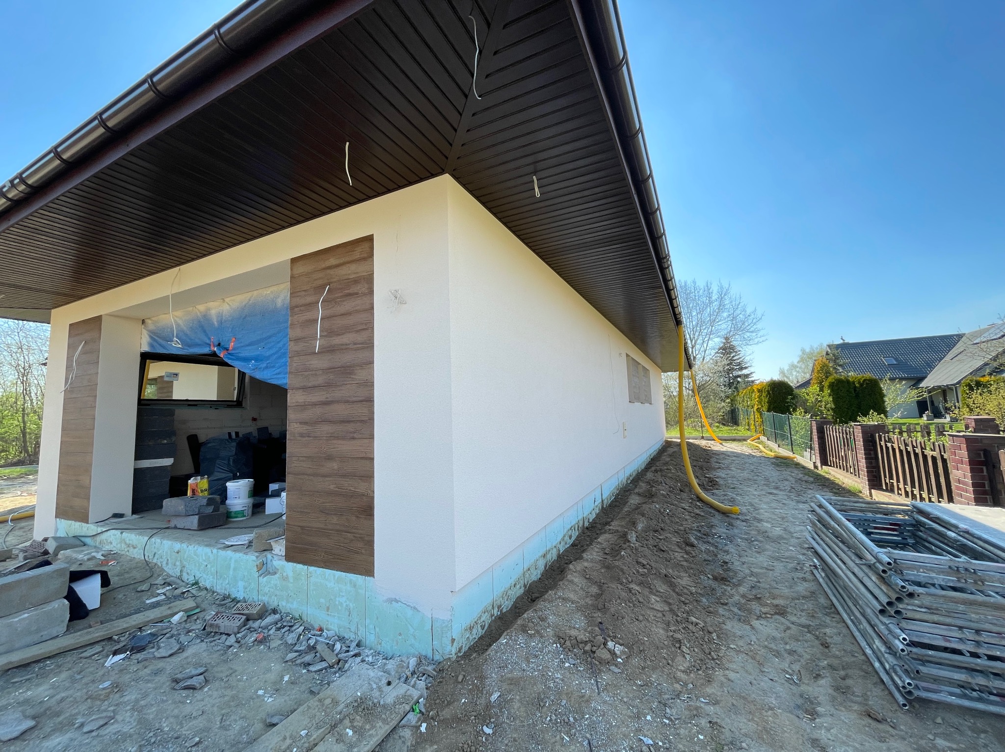 Budynek w trakcie prac elewacyjnych: ocieplona podstawa, częściowo wykończona ściana z dekoracyjnymi panelami drewnopodobnymi, widoczne rusztowania i materiały budowlane.