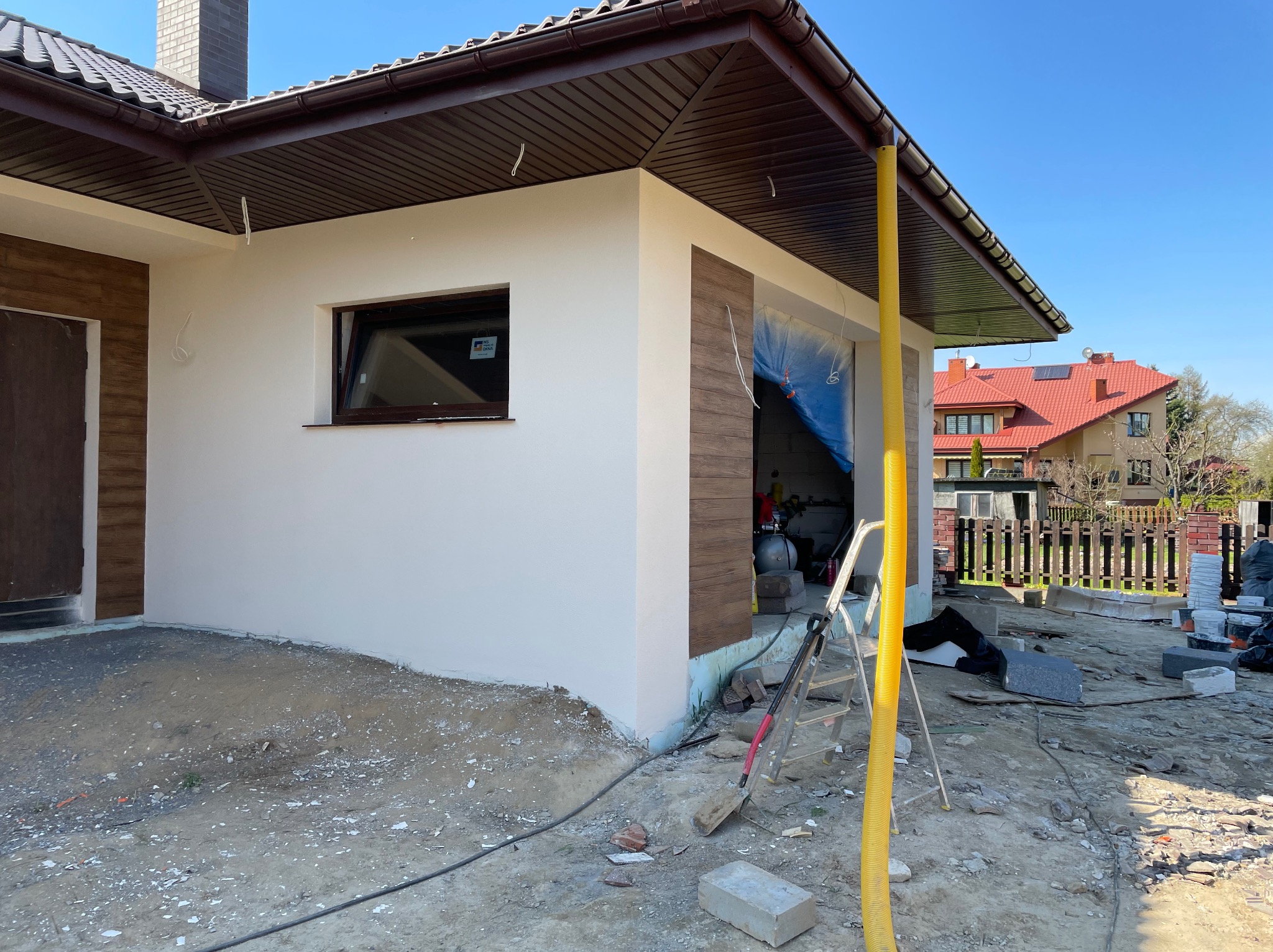 Świeżo otynkowana elewacja domu jednorodzinnego z brązowymi elementami dekoracyjnymi, widoczne narzędzia i materiały budowlane sugerujące trwające prace wykończeniowe.