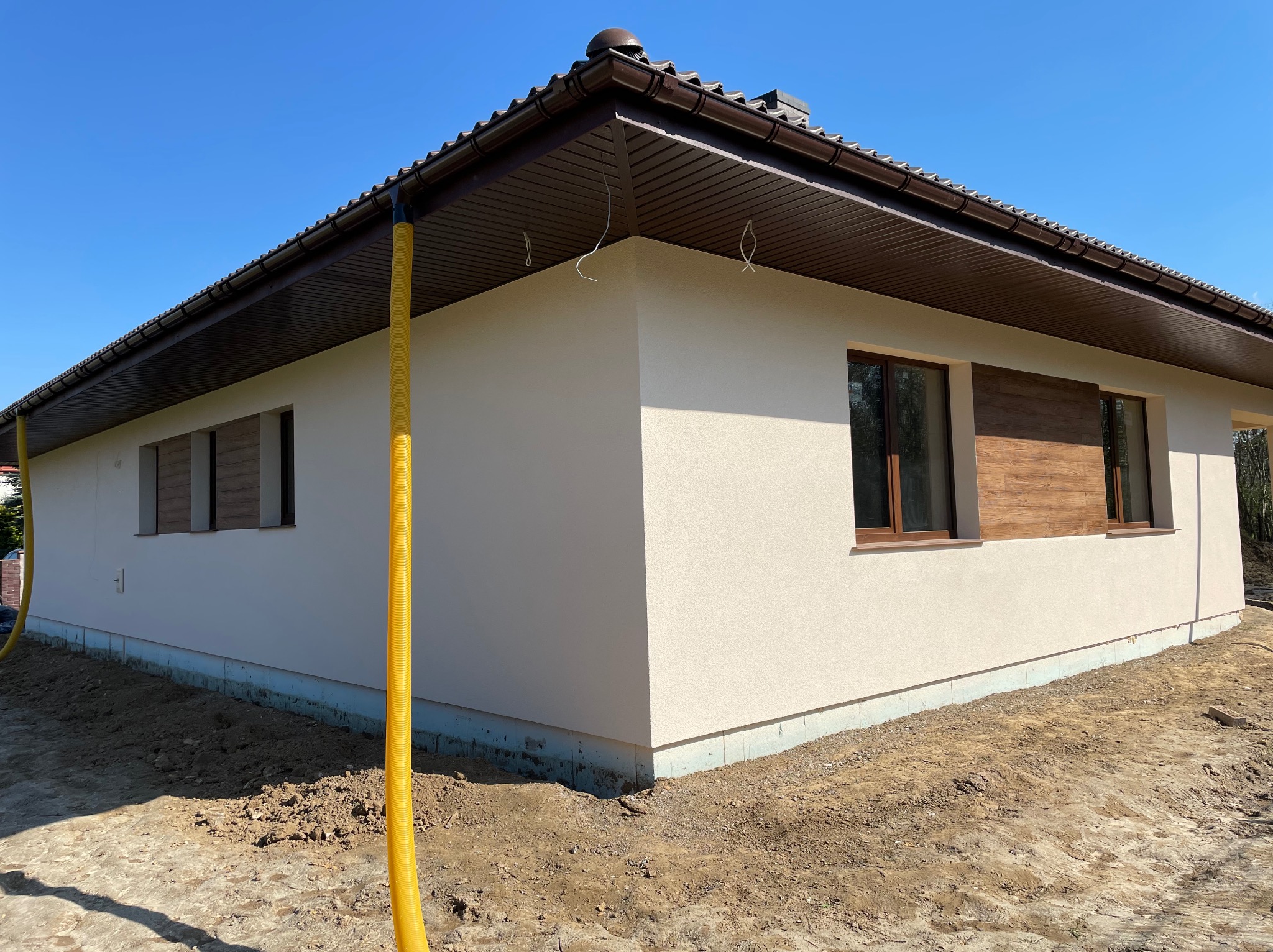 Nowoczesny dom jednorodzinny z jasną elewacją, brązową stolarką okienną i dachem, widoczne żółte rury wentylacyjne przy elewacji, teren wokół w trakcie prac ziemnych.