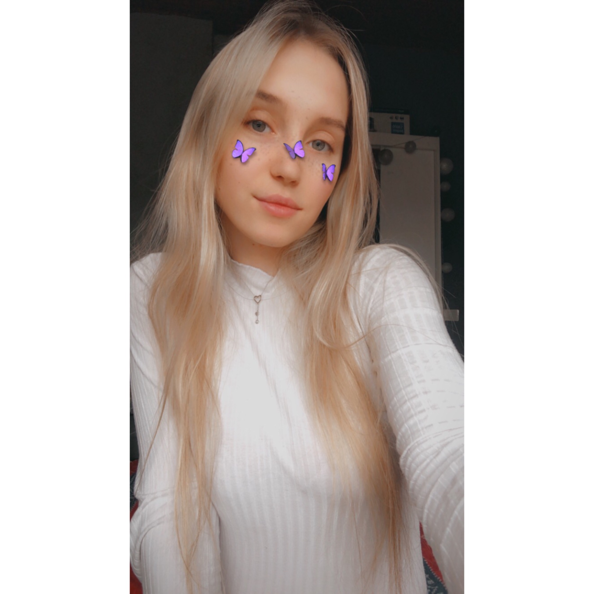 Portret młodej kobiety z długimi blond włosami, ozdobionymi fioletowymi motylkami na twarzy, w białej bluzce na tle ciemnego pokoju.