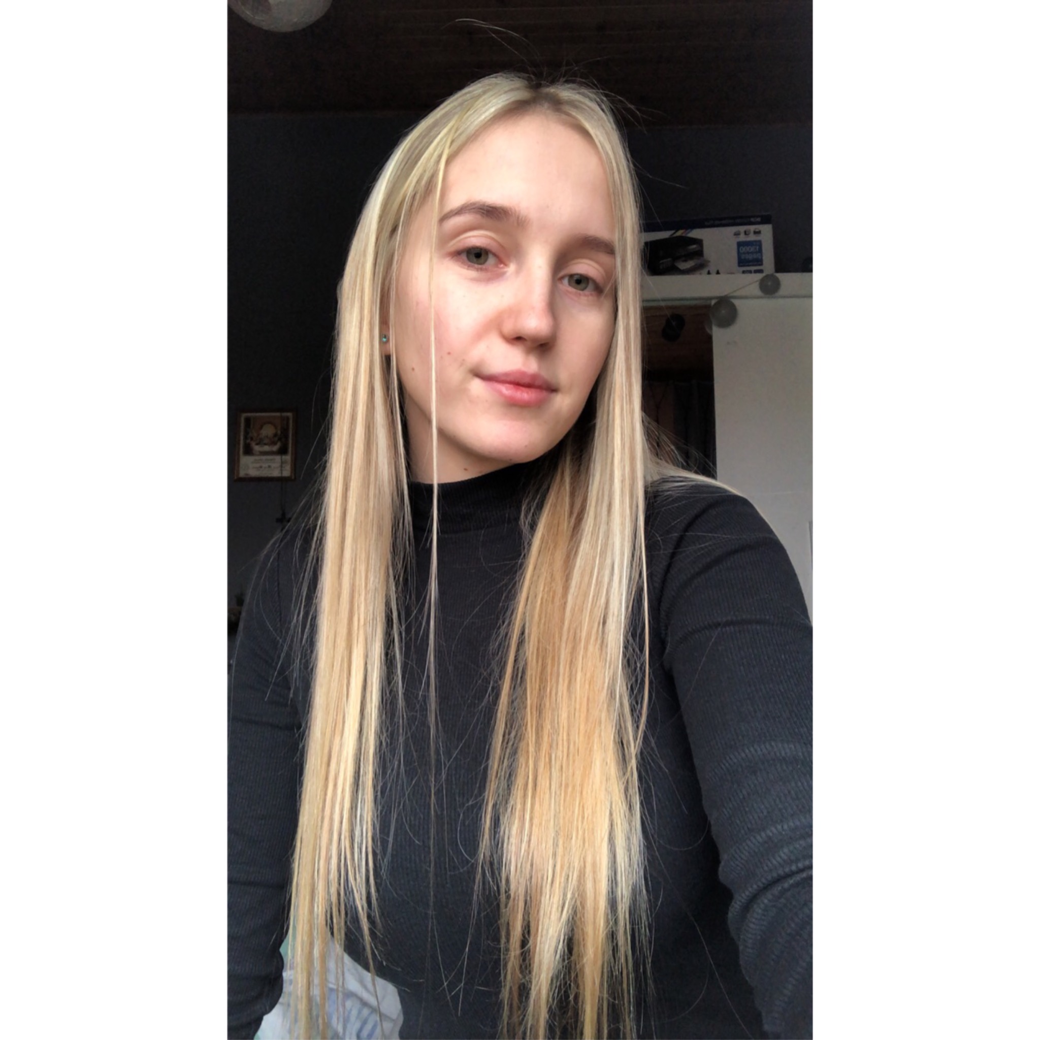 Portret młodej kobiety z długimi, prostymi blond włosami, ubrana w czarny golf, z delikatnym makijażem, pozująca w domowym wnętrzu z widoczną drukarką Brother w tle.