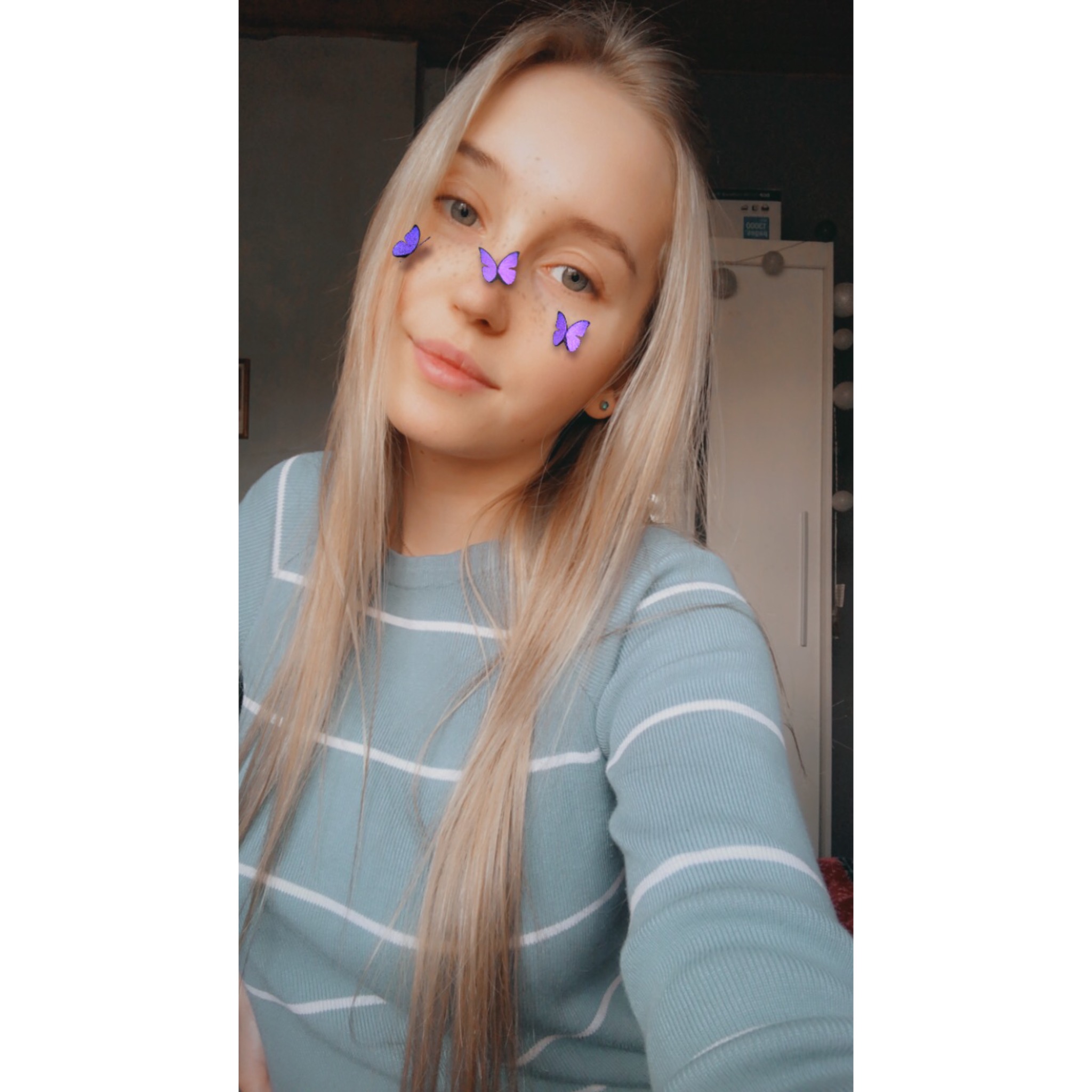 Portret uśmiechniętej młodej kobiety z blond włosami i efektem motyli na twarzy, w tle widoczna szafa.