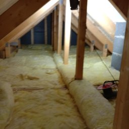 Extrem - Poddasze w trakcie ocieplania wełną mineralną, widoczne drewniane elementy konstrukcyjne dachu i ściany szczytowej, na podłodze rozłożona wełna, w tle czarna ściana działowa oraz zwinięty przedłużacz.