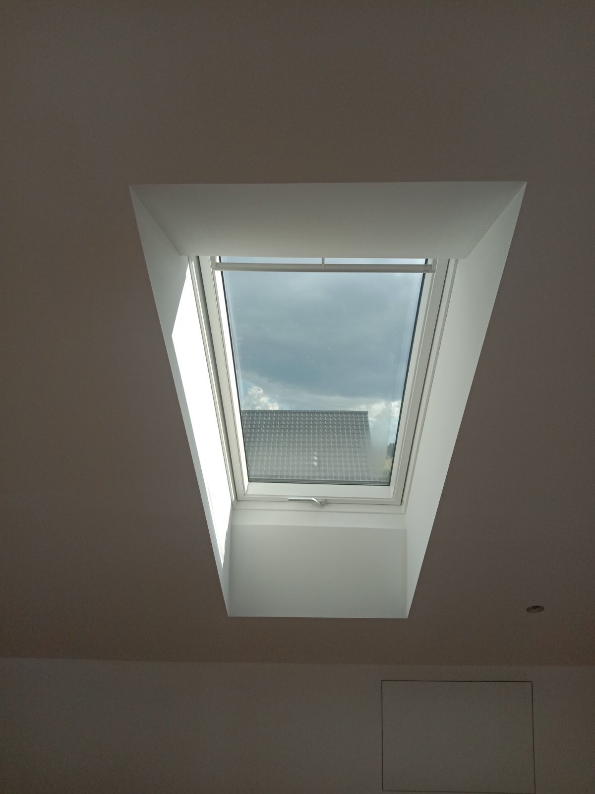 Białe okno dachowe w skośnym suficie, widok na pochmurne niebo i fragment dachu sąsiedniego budynku. Minimalistyczny design, jasne wnętrze.