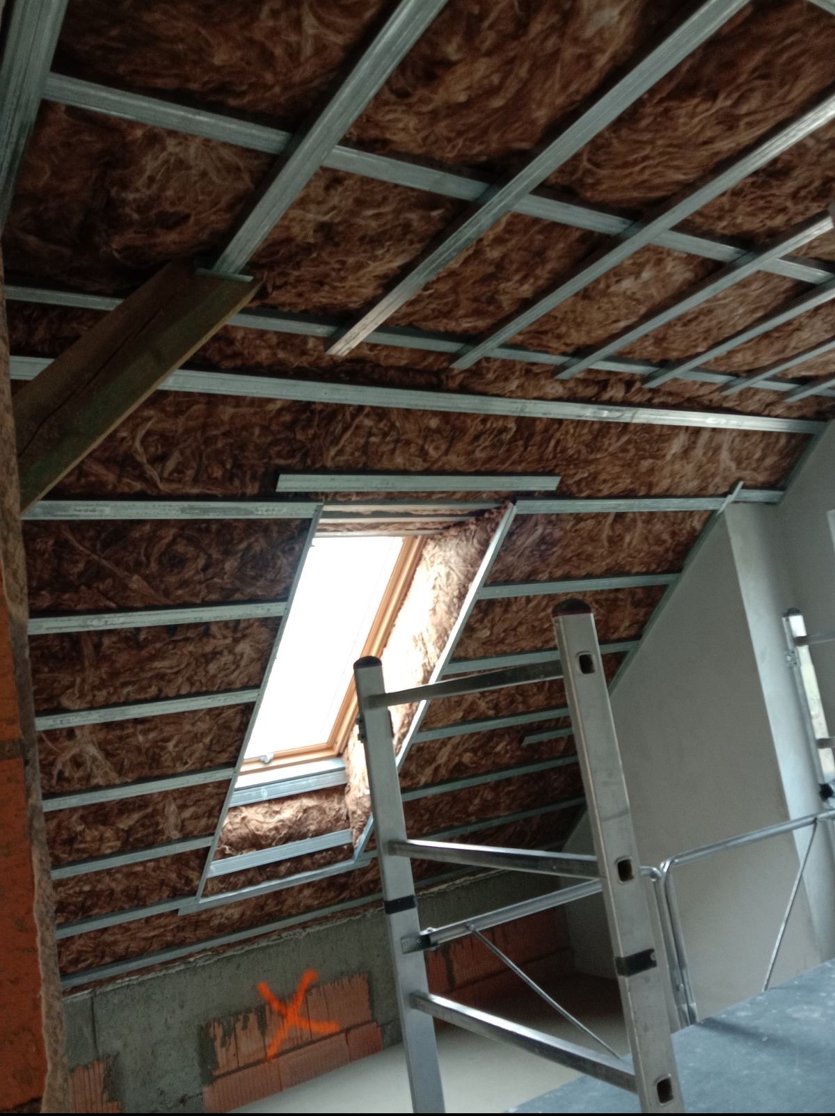 Skośny strych z oknem dachowym w trakcie izolacji wełną mineralną i montażu metalowego stelażu pod płyty gipsowo-kartonowe, widok z dołu na drabinę.