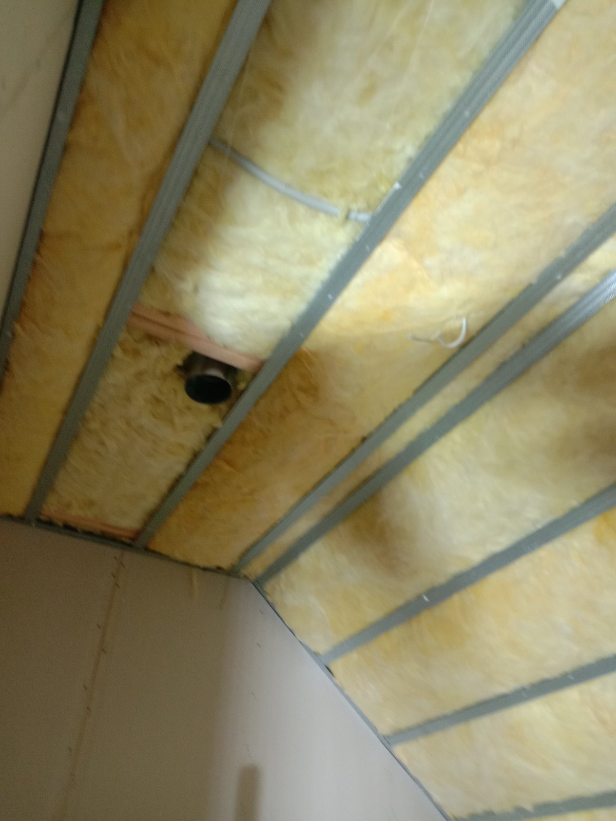 Montaż izolacji z wełny mineralnej w konstrukcji sufitu podwieszanego z profili metalowych, widoczny otwór wentylacyjny.