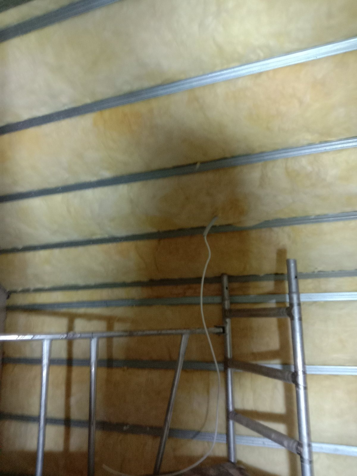 Montaż izolacji z wełny mineralnej na suficie podwieszanym, widoczne metalowe profile i fragment rusztowania.