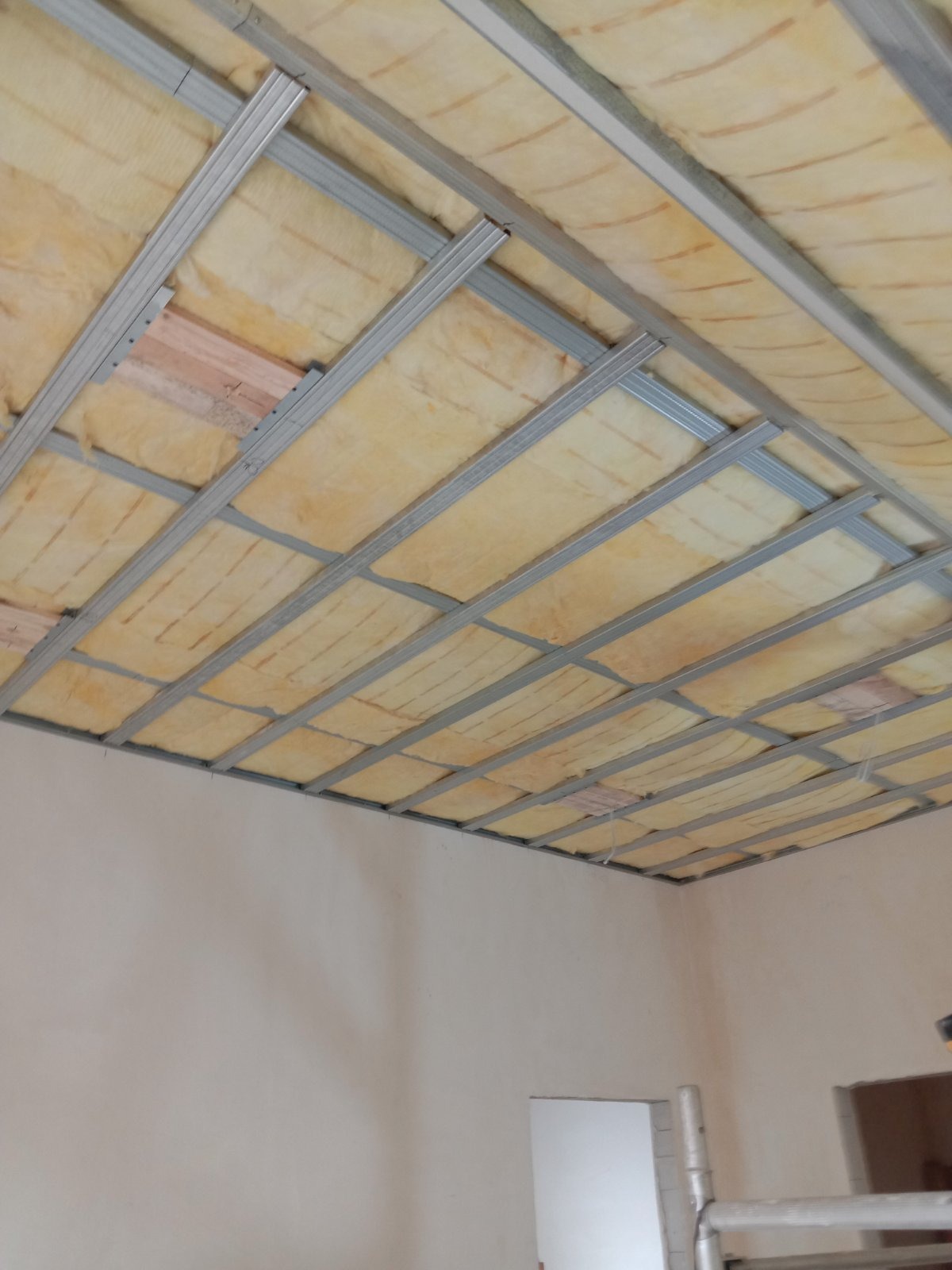 Montaż sufitu podwieszanego: widoczna metalowa konstrukcja nośna wypełniona wełną mineralną, przygotowana do dalszych prac wykończeniowych w pomieszczeniu z pomalowanymi ścianami.