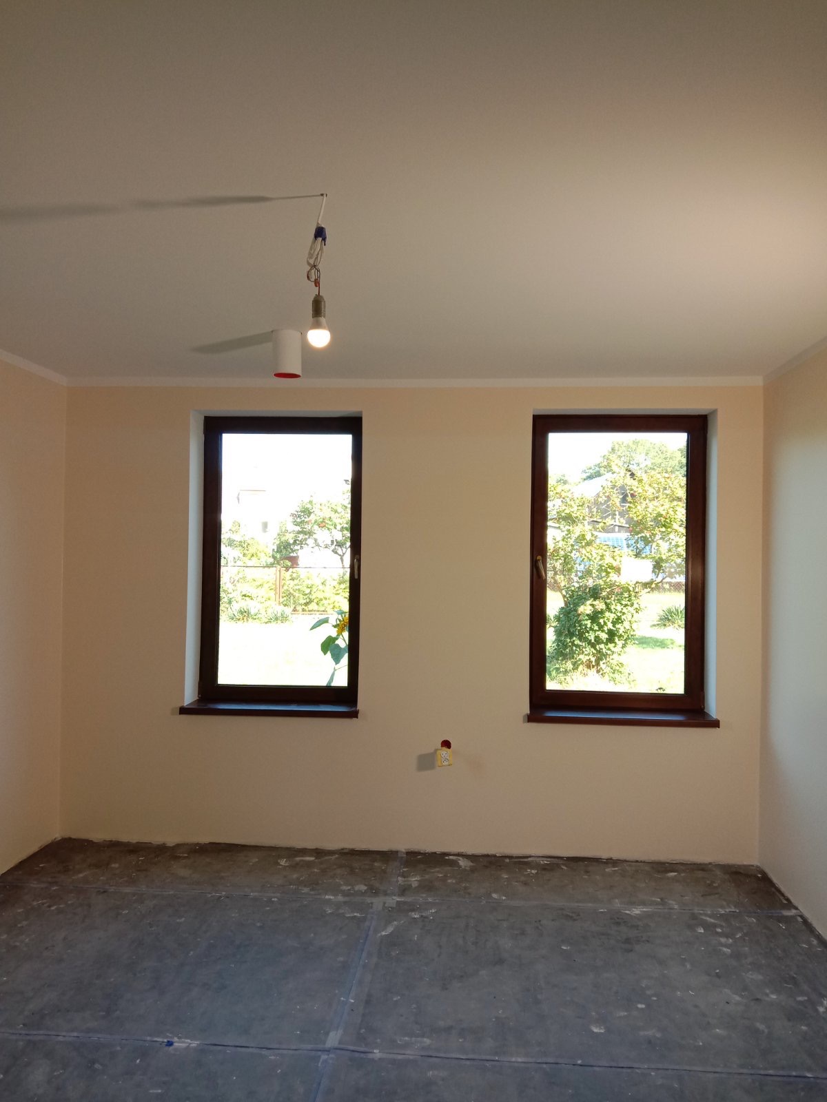 Pokój w trakcie prac przygotowawczych: ściany pomalowane na jasny kolor, sufit wykończony, podłoga przygotowana pod dalsze prace, widoczne dwa okna z drewnianymi ramami i tymczasowe oświetlenie.