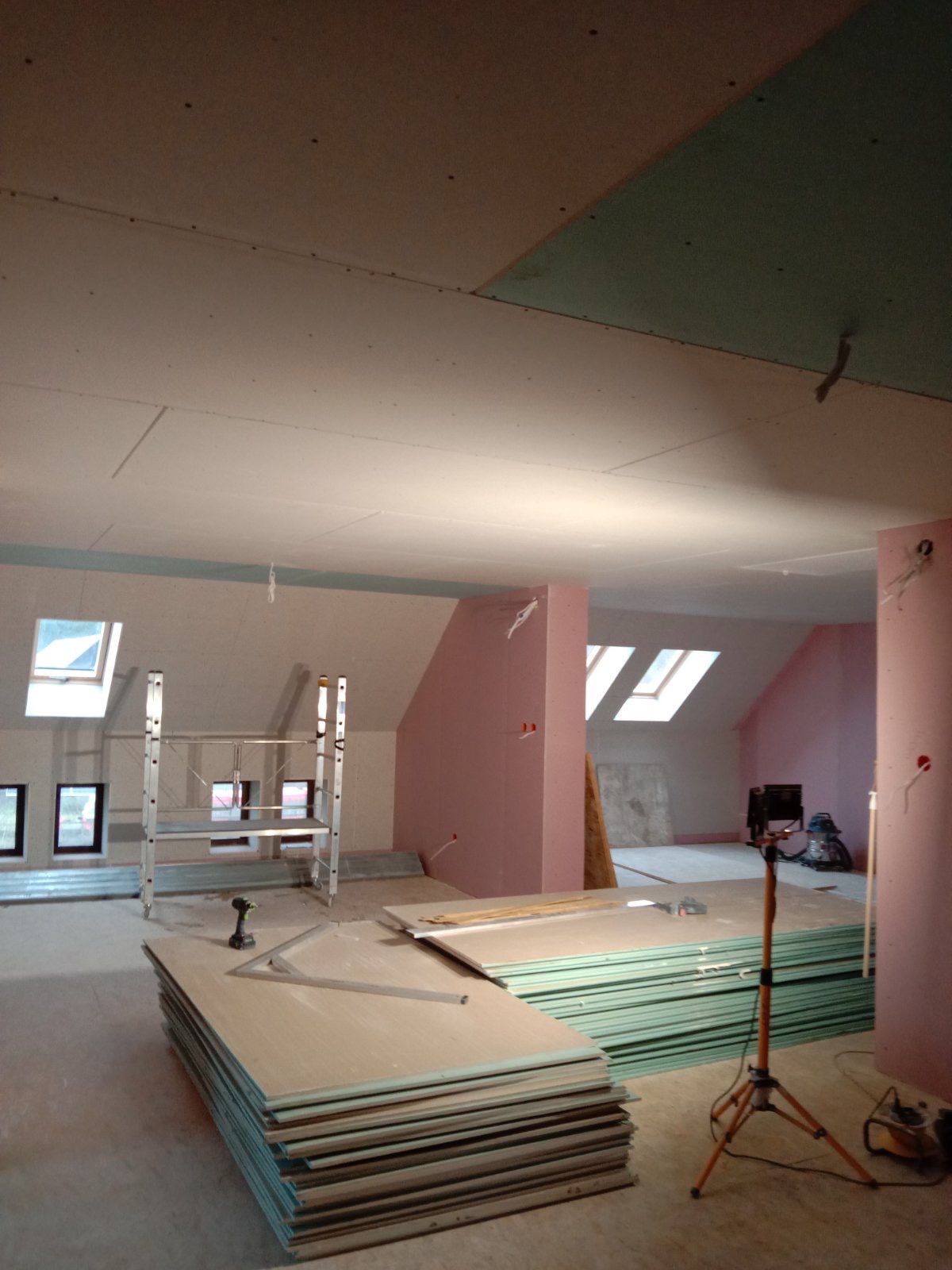 Poddasze w trakcie adaptacji: stelaż, płyty gipsowo-kartonowe ułożone na podłodze, widoczne okna dachowe i ściany ocieplone wełną mineralną w kolorze różowym, lampa budowlana na trójnogu.