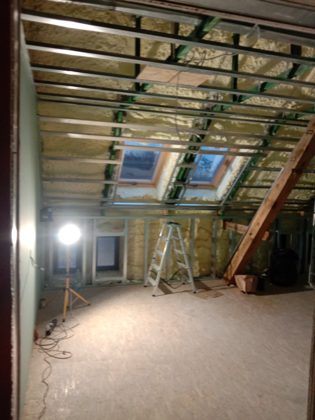 Poddasze w trakcie ocieplania pianą PUR, widoczne okna dachowe, metalowa konstrukcja sufitu, drabina i lampa halogenowa na statywie.