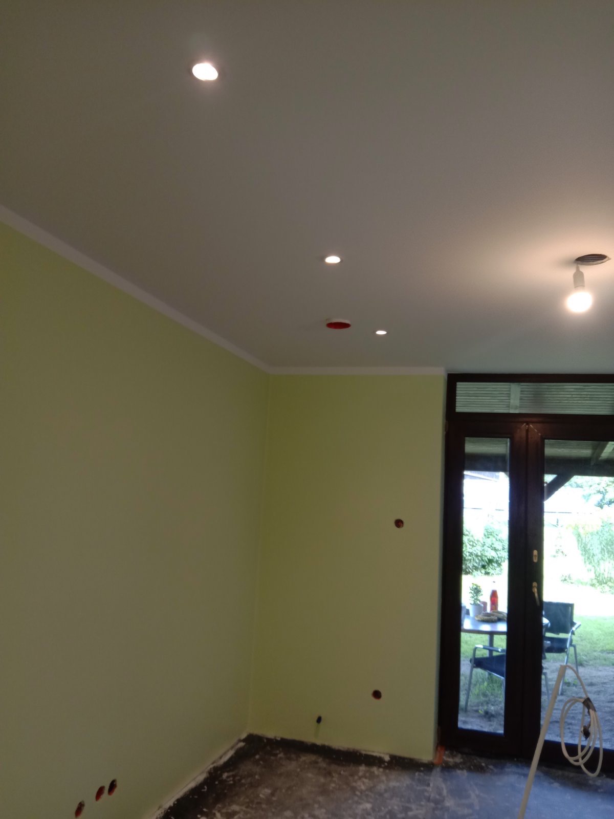 Wnętrze pokoju w trakcie prac wykończeniowych: świeżo pomalowane ściany w odcieniach zieleni, biały sufit z zamontowanymi punktowymi lampami LED, widoczne otwory na gniazdka elektryczne w ścianach,...