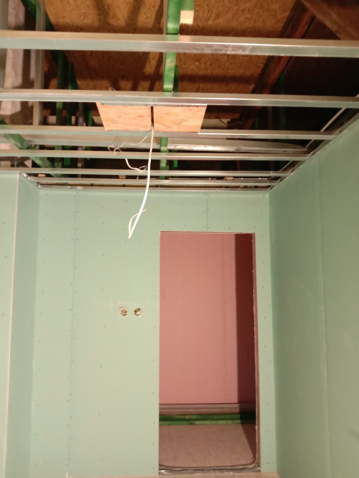 Wnętrze w trakcie prac wykończeniowych: ściany obłożone płytami gipsowo-kartonowymi w kolorze zielonym, widoczna metalowa konstrukcja sufitu podwieszanego z wystającymi przewodami elektrycznymi...