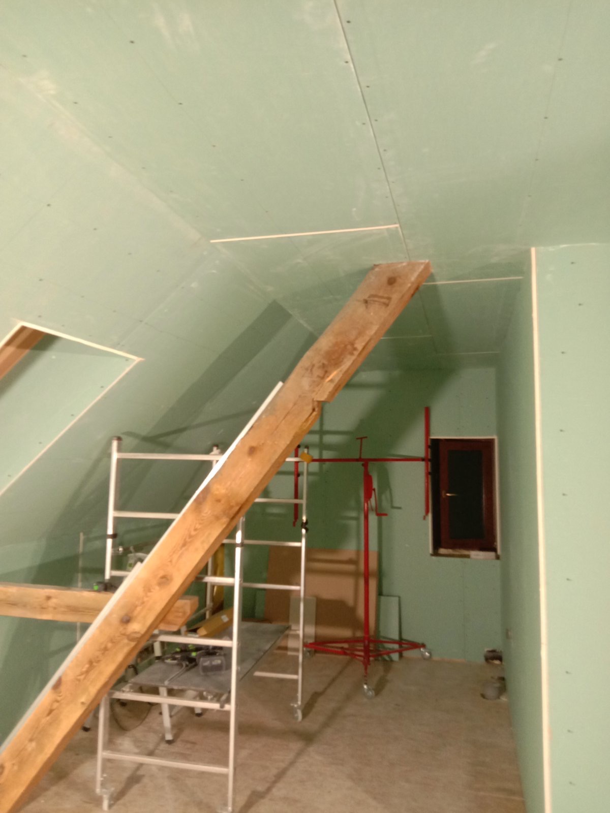 Poddasze w trakcie prac wykończeniowych: widoczne płyty gipsowo-kartonowe na ścianach i skośnym suficie, drewniane elementy konstrukcyjne, rusztowanie oraz podnośnik do płyt, okno w brązowej ramie.