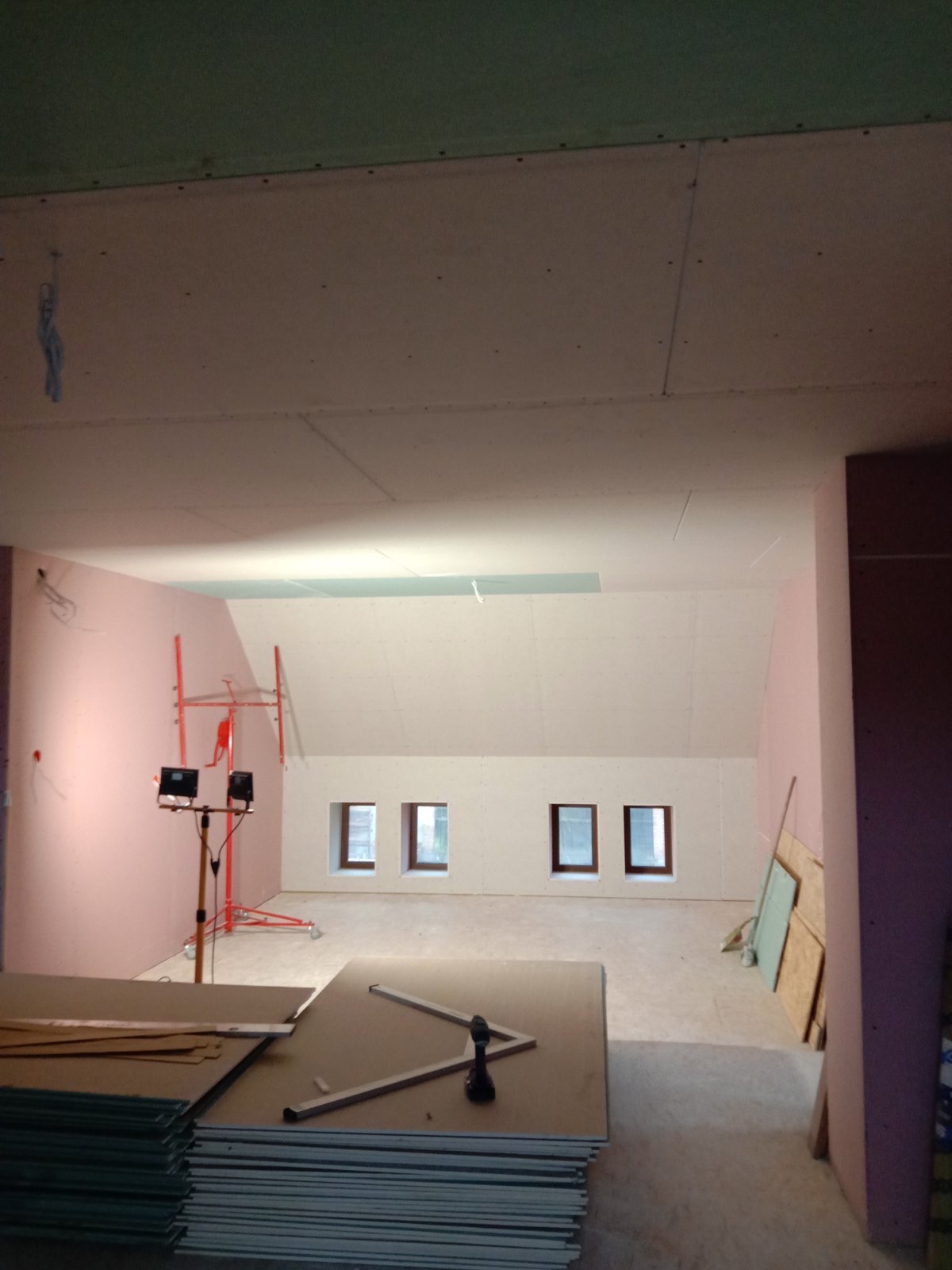 Poddasze w trakcie prac wykończeniowych: ściany i sufit obłożone płytami gipsowo-kartonowymi, widoczne narzędzia i materiały budowlane, oświetlenie robocze, trzy okna dachowe.