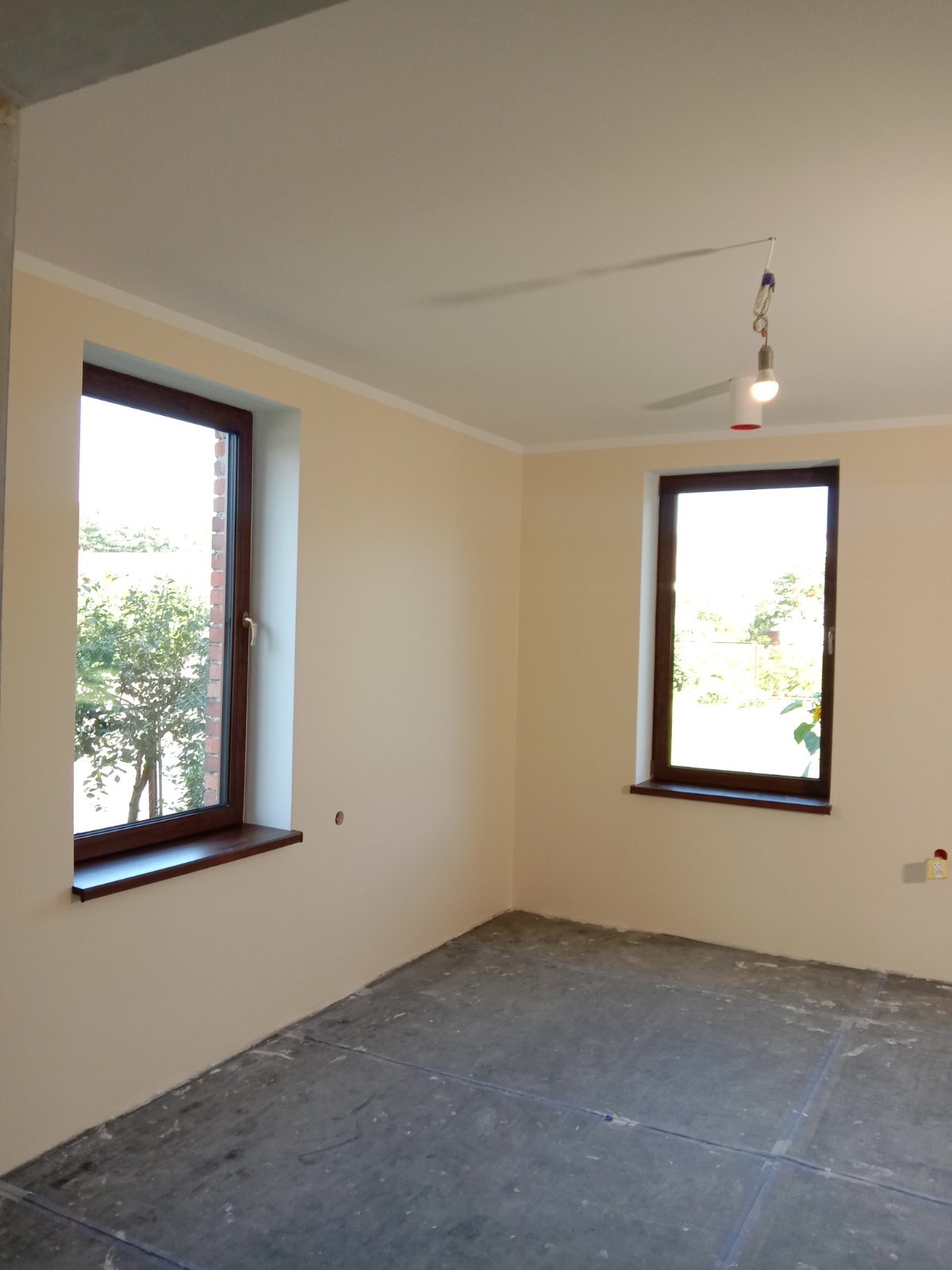 Wnętrze pokoju po świeżym malowaniu ścian na jasny odcień, widoczne dwa okna z ciemnymi ramami i surowa podłoga przygotowana do dalszych prac wykończeniowych w Goleniowie, tymczasowe oświetlenie...