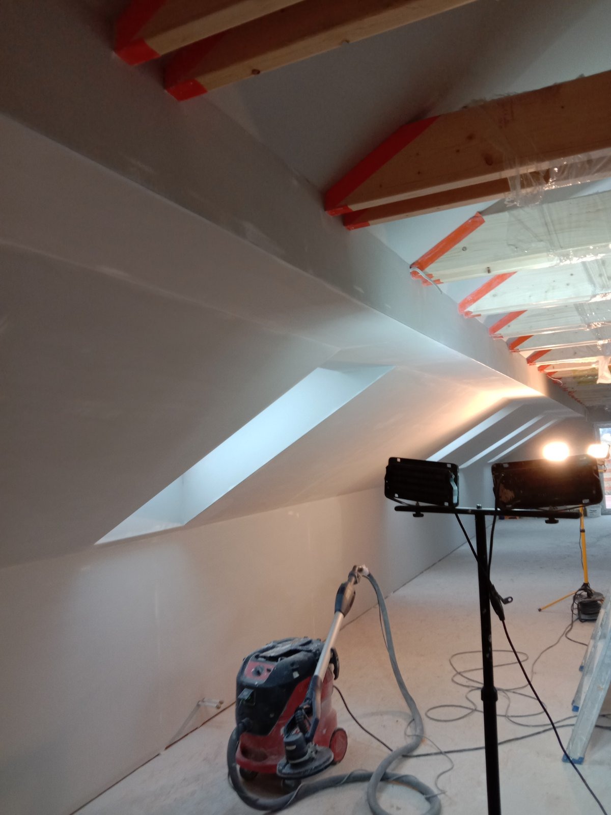 Poddasze po nałożeniu gładzi, widoczne okno dachowe, szlifierka do gładzi z odkurzaczem, oświetlenie budowlane i zabezpieczone elementy drewniane.