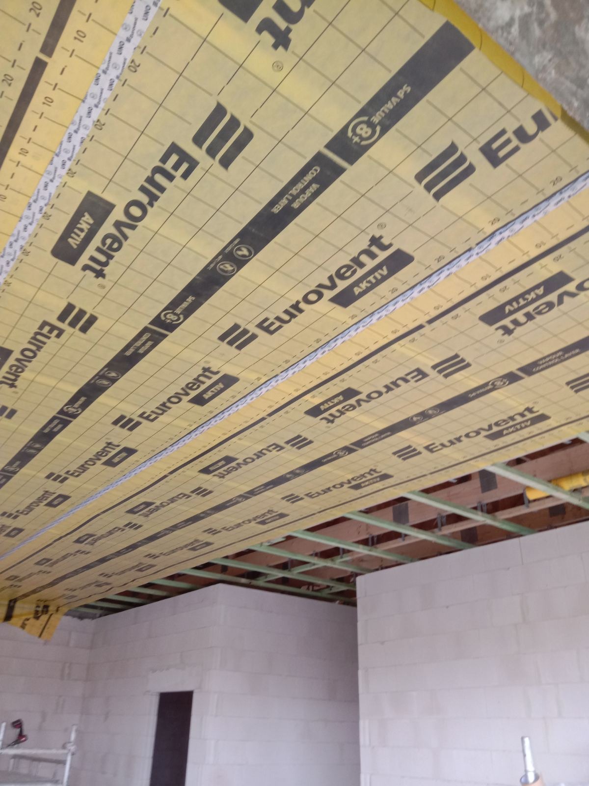 Żółta folia paroizolacyjna Eurovent Aktiv, z nadrukowaną siatką i logo, rozciągnięta na drewnianym ruszcie sufitowym w surowym stanie, widoczne ściany z bloczków i fragment rusztowania.