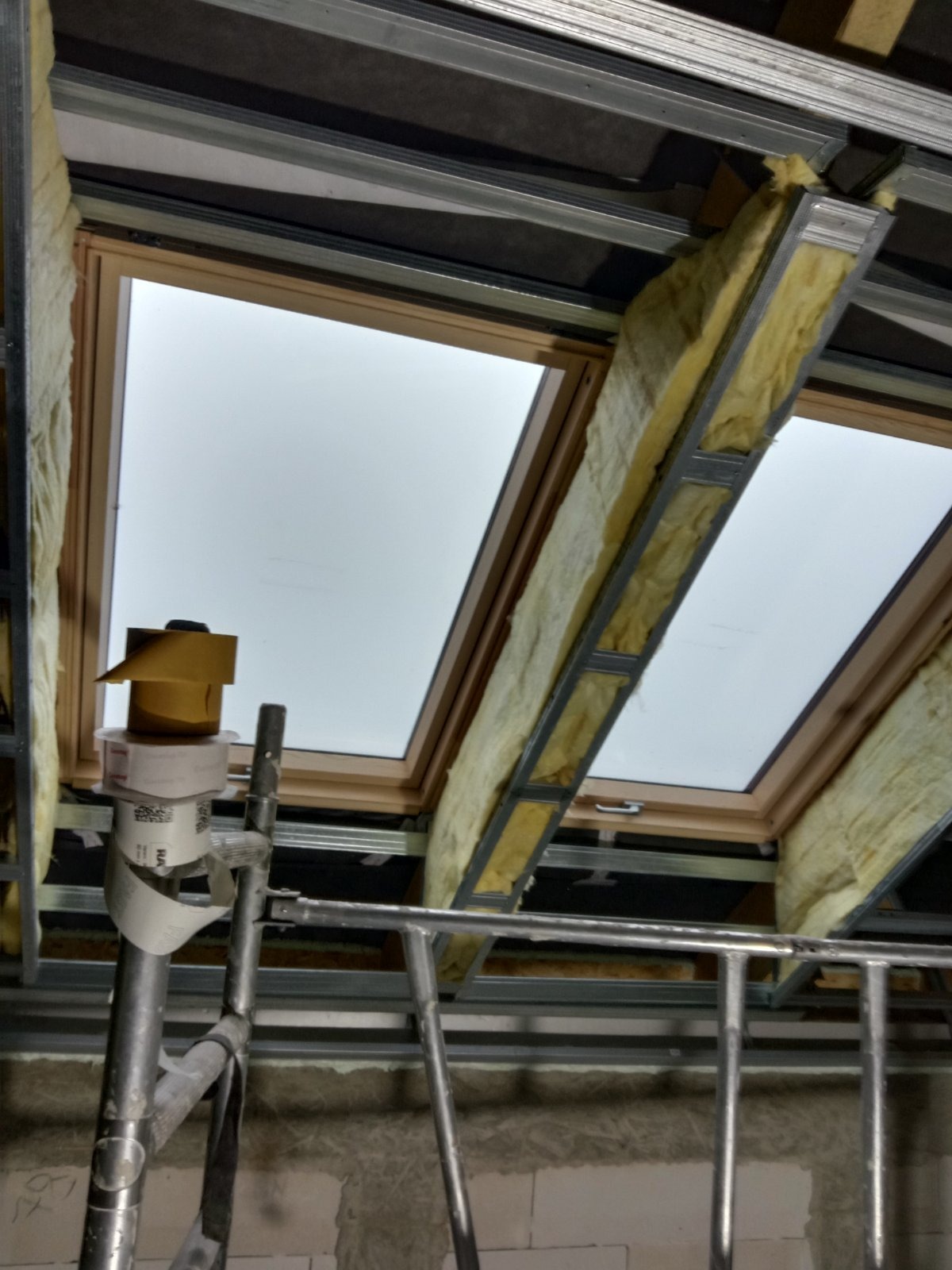 Montaż okien dachowych z izolacją wełną mineralną, widoczne metalowe profile i drabina malarska w surowym wnętrzu.