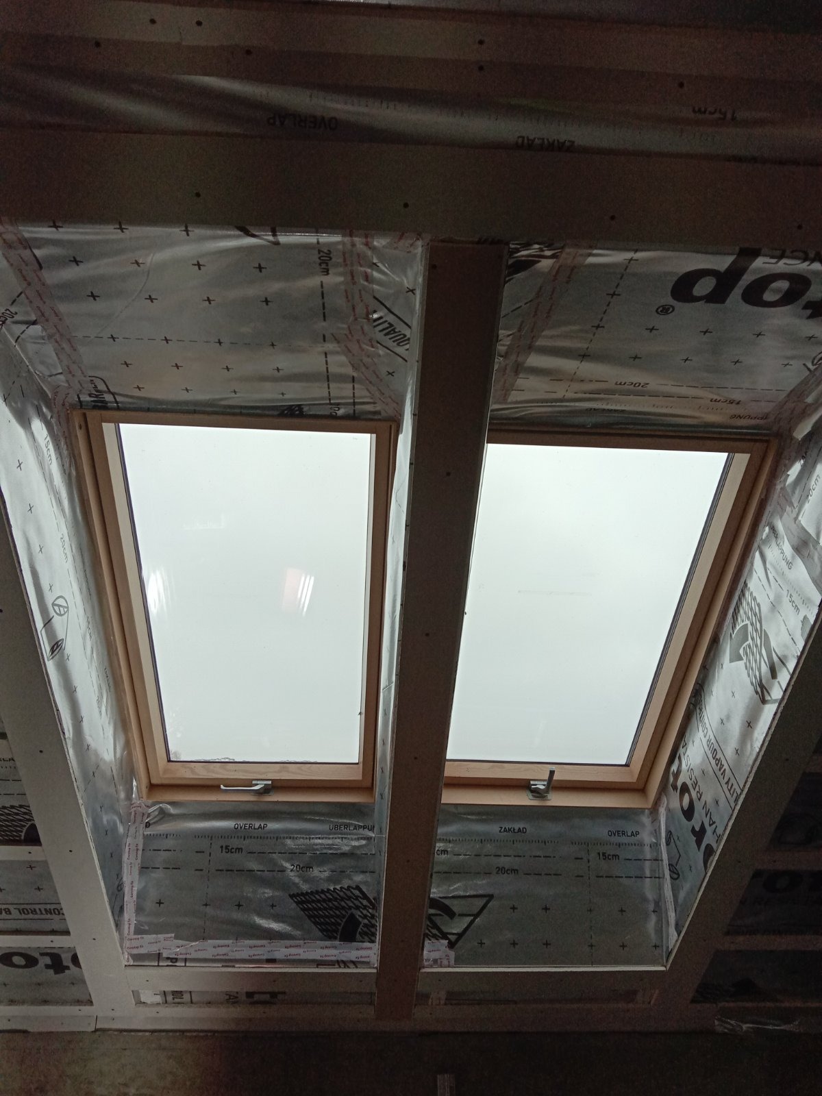 Montaż dwóch okien dachowych w drewnianej konstrukcji, widoczne folie izolacyjne z nadrukami, surowe drewno, widok od dołu.