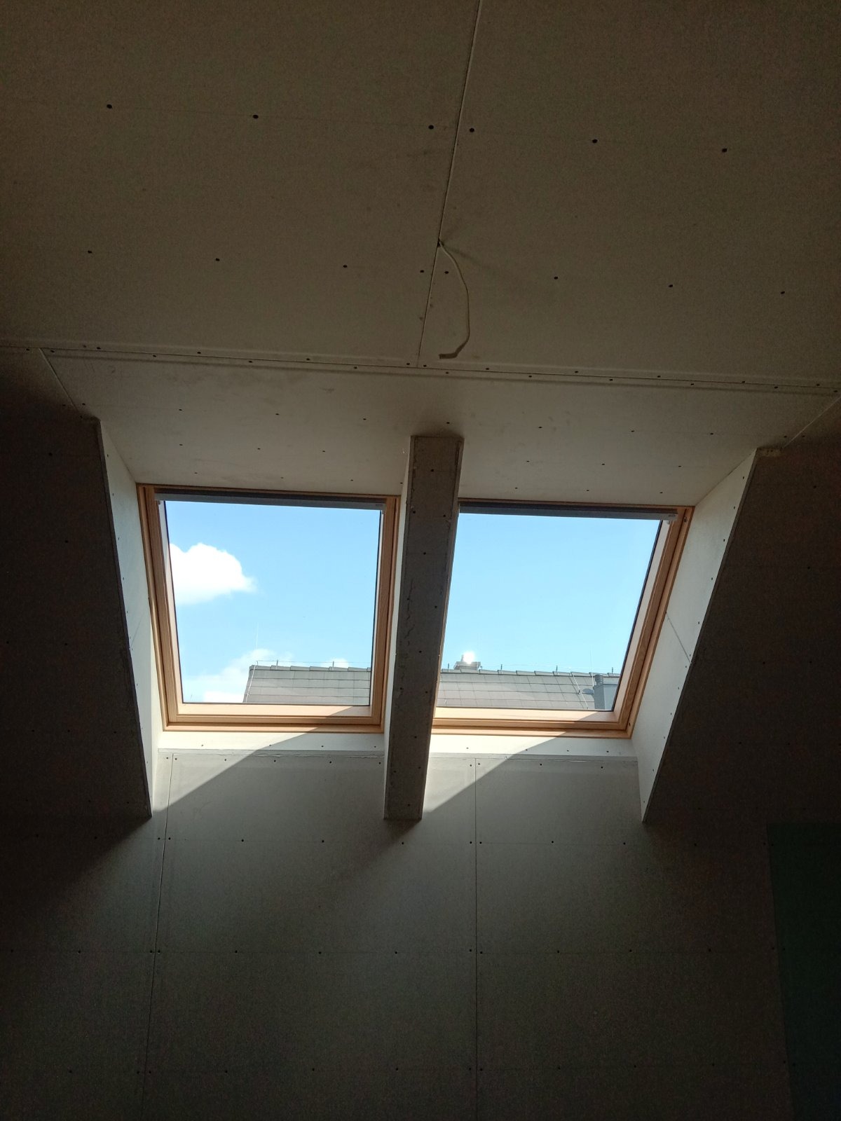 Dwa drewniane okna dachowe połączone słupkiem, widok na jasne niebo i fragment dachu sąsiedniego budynku, ściany i sufit pokryte szarą płytą gipsowo-kartonową przed wykończeniem.