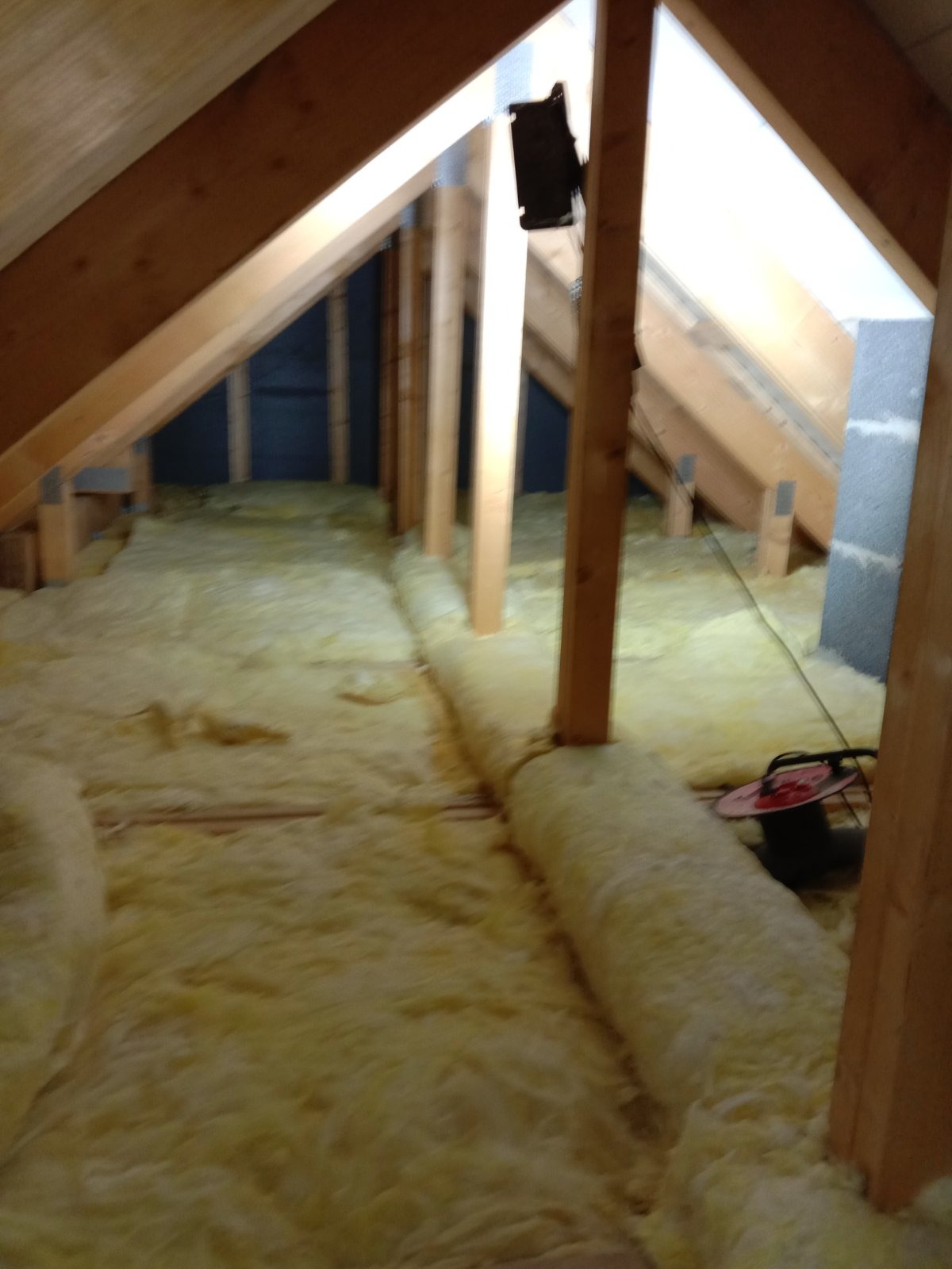 Poddasze w trakcie ocieplania wełną mineralną, widoczne drewniane elementy konstrukcyjne dachu i ściany szczytowej, na podłodze rozłożona wełna, w tle czarna ściana działowa oraz zwinięty przedłużacz.