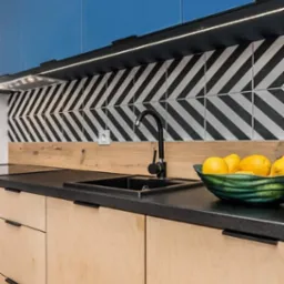 Nowoczesna kuchnia z jasnego drewna, czarnym blatem i baterią, biało-czarne płytki w jodełkę na ścianie, oświetlenie LED pod szafkami, misa z cytrynami.