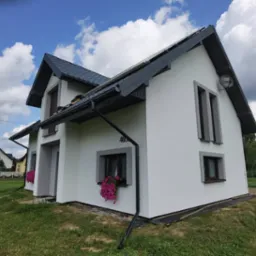 Dwukondygnacyjny dom jednorodzinny z białą elewacją, szarą stolarką okienną i ciemnoszarym dachem, z numerem 403 i anteną satelitarną na ścianie, z kwiatami w skrzynkach pod oknami.