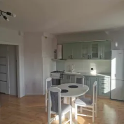 Jasna kuchnia z drewnianą podłogą, okrągłym stołem i pastelowymi szafkami. W tle widoczny fragment przedpokoju z drzwiami.