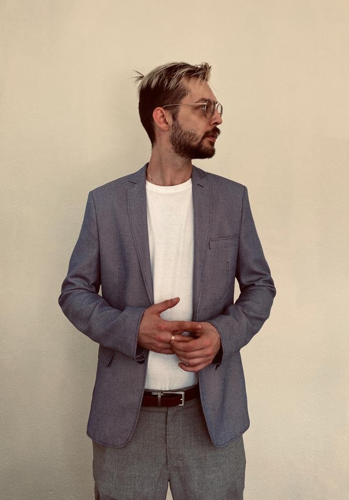 Mężczyzna w okularach, jasnej koszulce i marynarce, z rękami splecionymi na wysokości brzucha, patrzy w bok na jasnym tle.