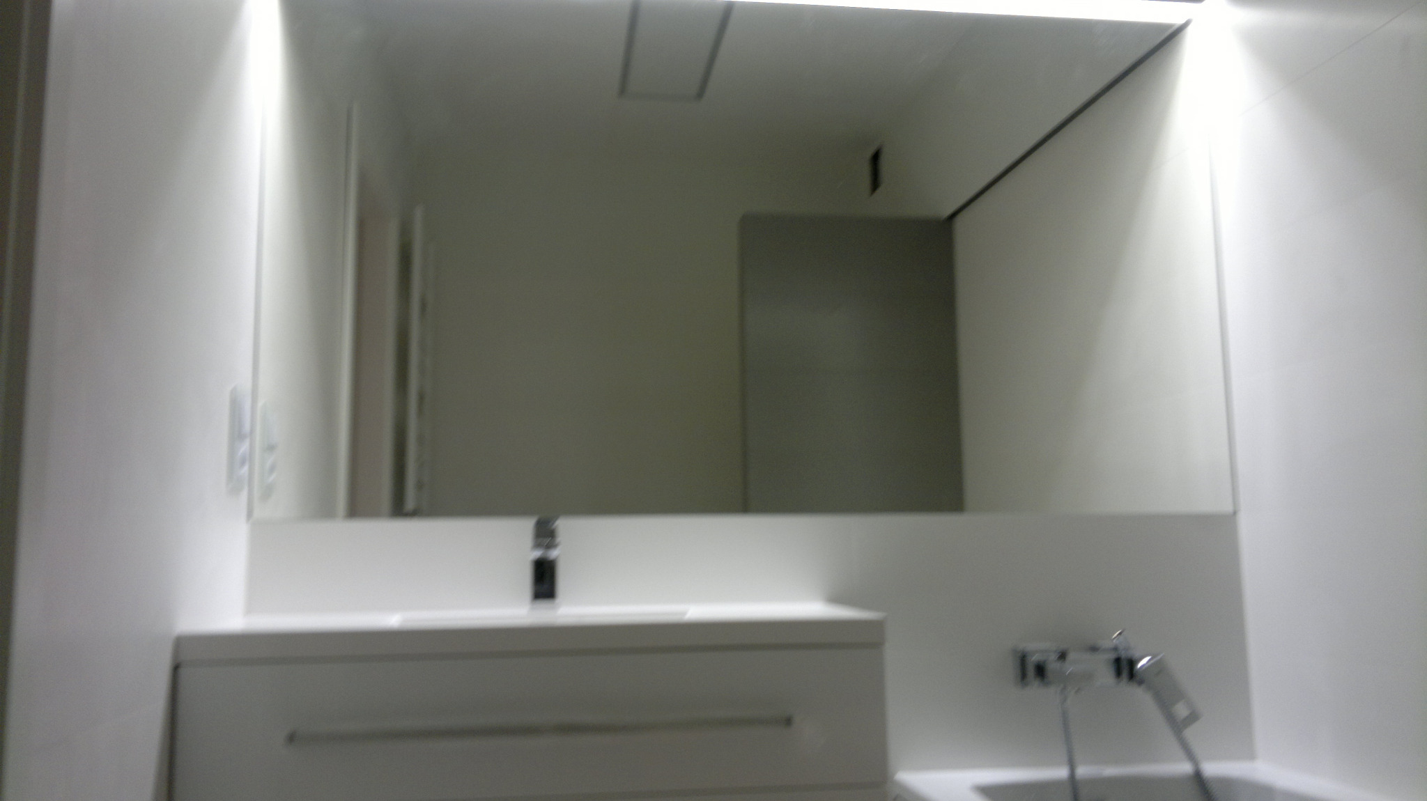 Minimalistyczna łazienka w bieli: prostokątne lustro nad umywalką z szafką, bateria umywalkowa w kolorze chromu, widoczny fragment baterii wannowej.