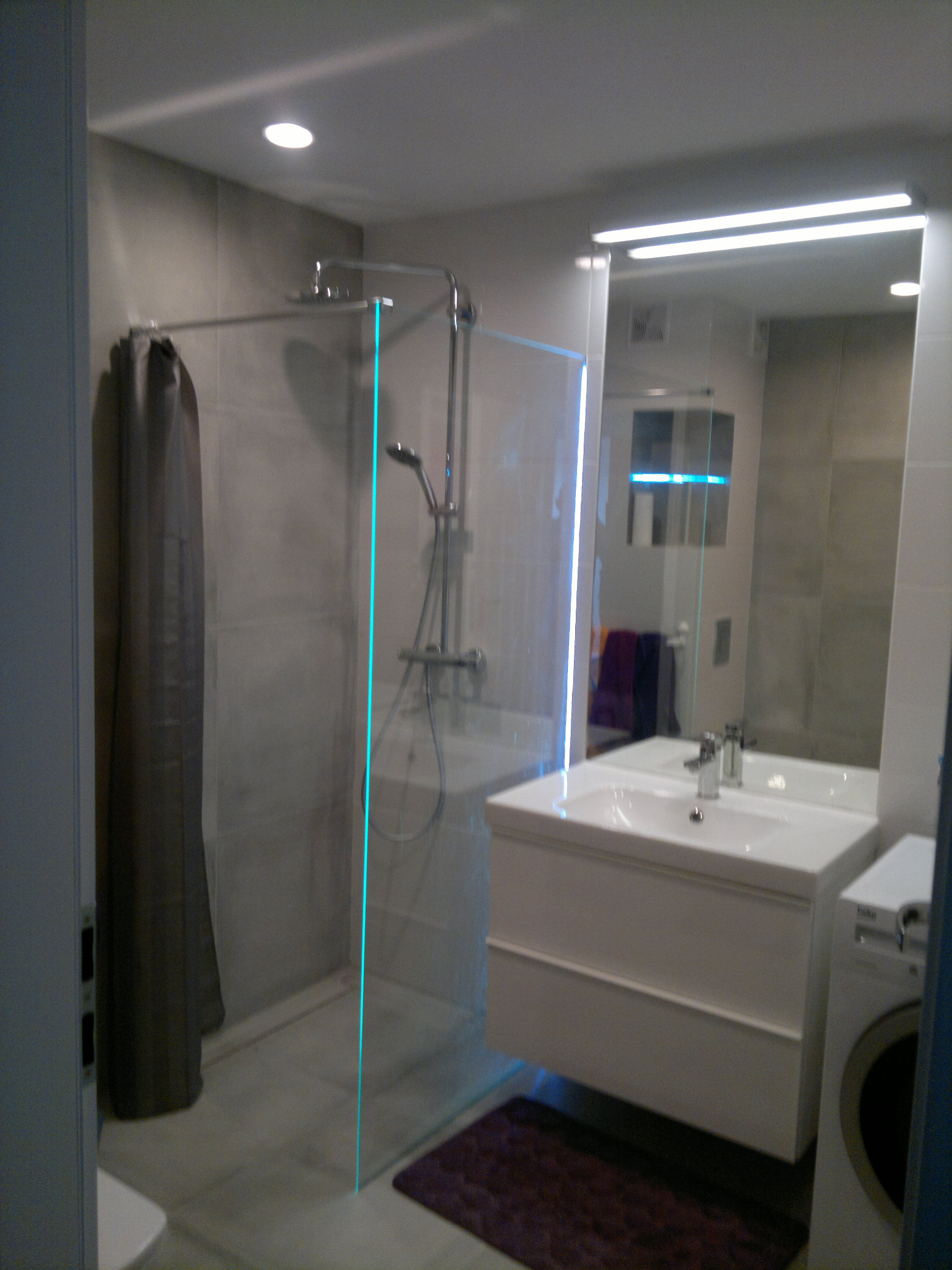 Nowoczesna łazienka z kabiną prysznicową typu walk-in z szarą zasłoną, białą szafką z umywalką, dużym lustrem z oświetleniem LED i pralką w rogu, wykończona szarymi płytkami.