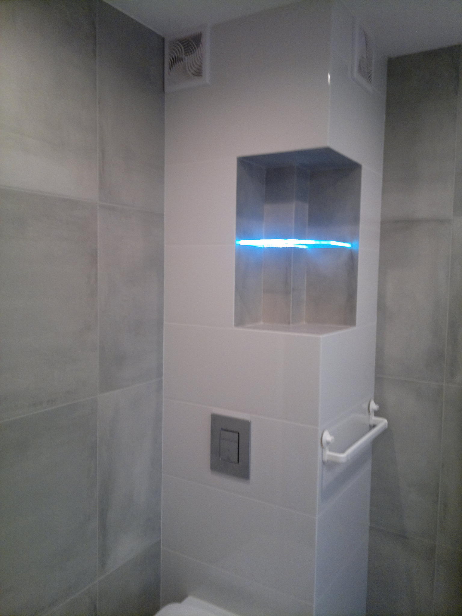 Nowoczesna łazienka z szarymi płytkami, podświetlaną wnęką w zabudowie WC, kwadratowym przyciskiem spłuczki i białym wieszakiem na ręczniki.