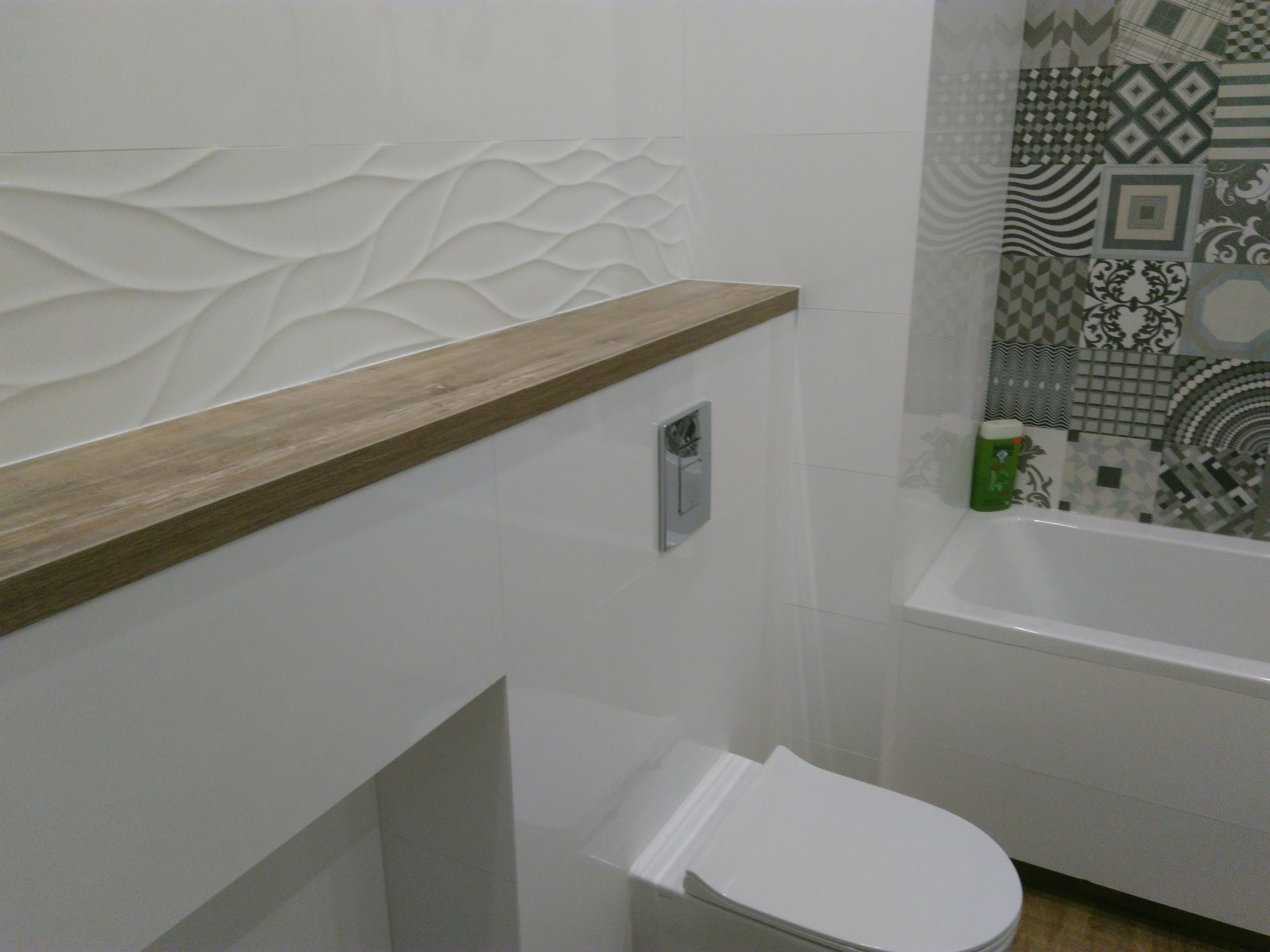 Nowoczesna łazienka z płytkami 3D na ścianie, drewnianą półką nad zabudowaną spłuczką WC i patchworkową mozaiką przy wannie.