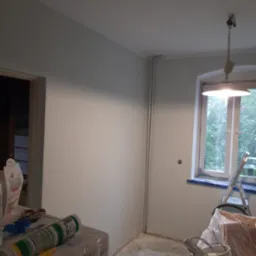 Świeżo pomalowane na jasno pomieszczenie z widocznymi narzędziami malarskimi, drabiną, lampą wiszącą i oknem z widokiem na zieleń.