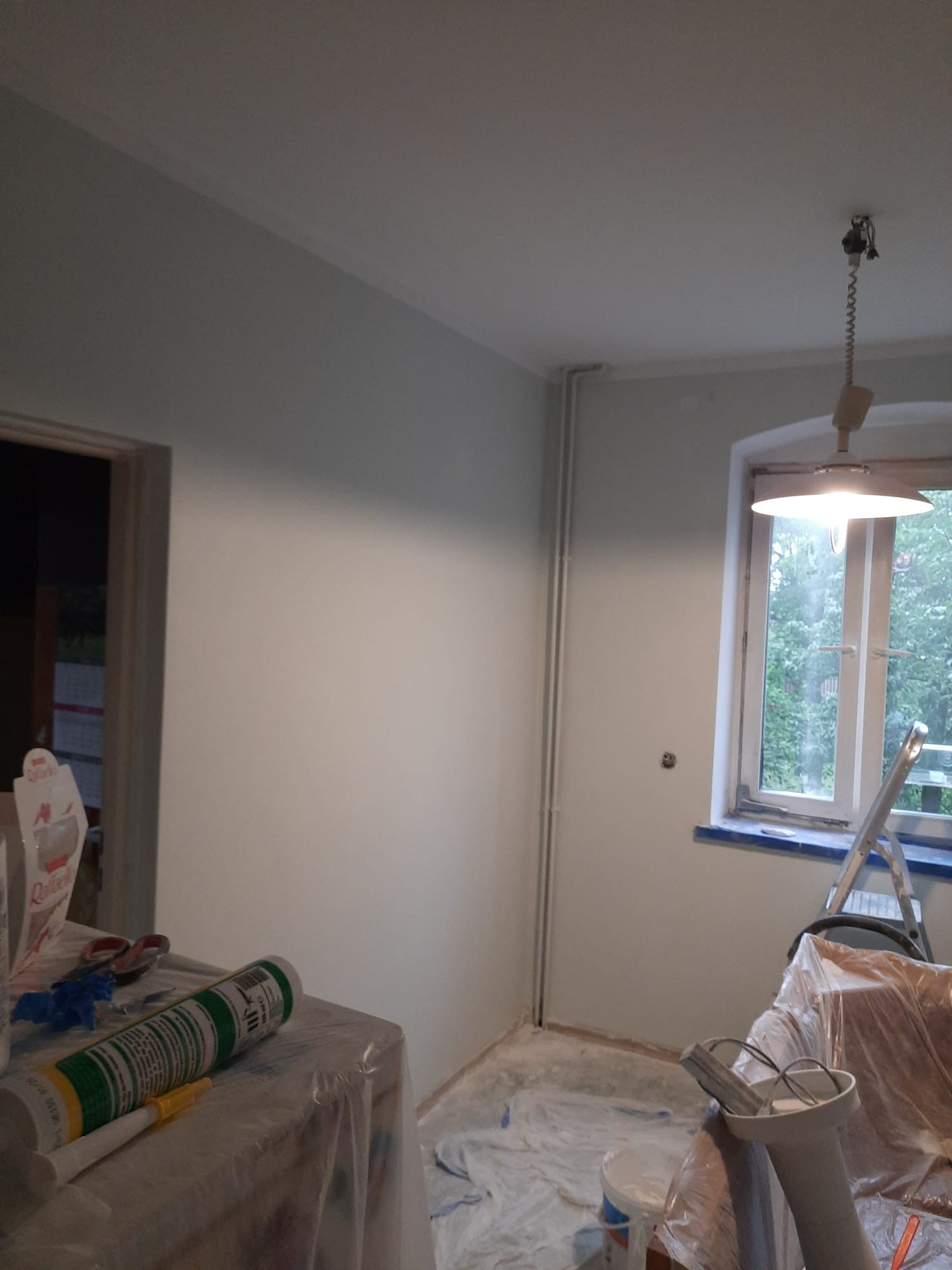 Świeżo pomalowane na jasno pomieszczenie z widocznymi narzędziami malarskimi, drabiną, lampą wiszącą i oknem z widokiem na zieleń.