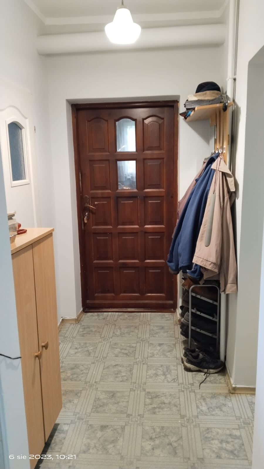 Wnętrze przedpokoju z drewnianymi drzwiami, szafką i wieszakiem na ubrania z półką na czapki oraz stojakiem na buty; podłoga wyłożona płytkami w kratkę.