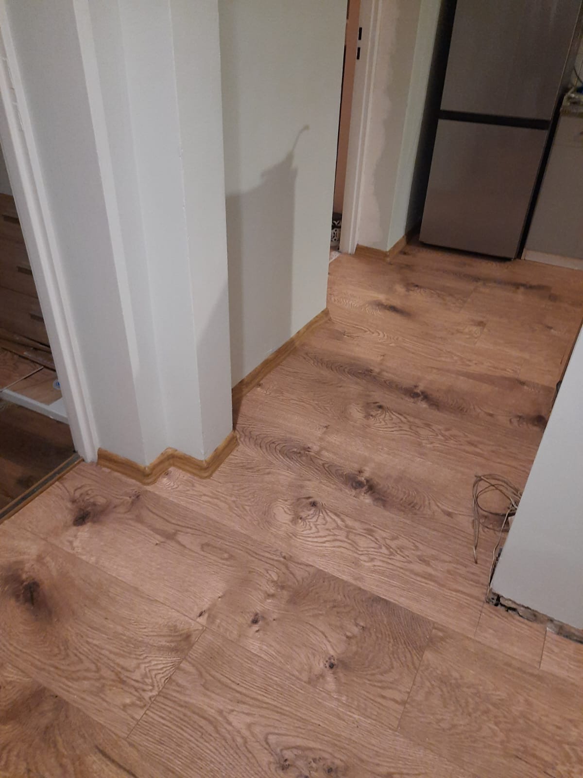 Ułożona podłoga z paneli drewnopodobnych w rogu pomieszczenia, widoczna listwa przypodłogowa i fragment ściany z kolumną oraz kable wystające ze ściany.