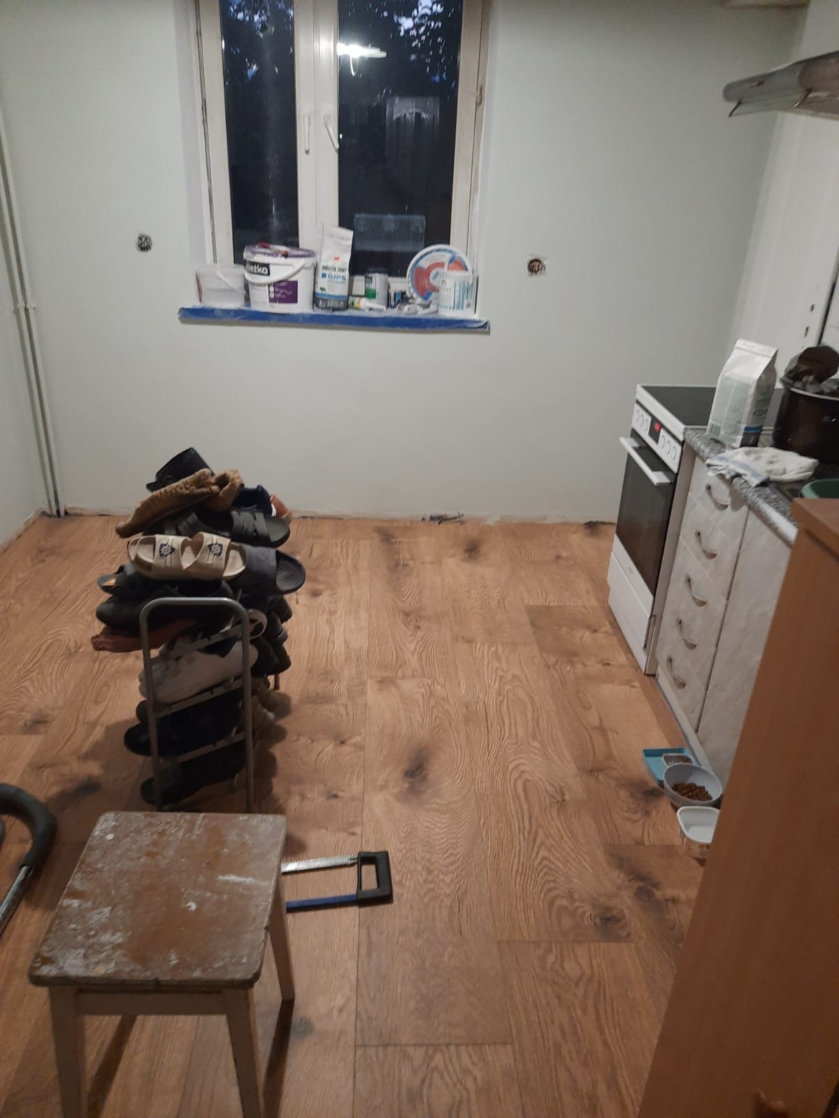 Nowa drewniana podłoga w kuchni podczas remontu, widoczny stos butów na metalowym stojaku, taboret i piła ręczna, okno z farbami i gładzią na parapecie, ślady po gniazdkach elektrycznych na ścianie.