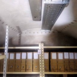 Michał Pawlusiak - Wilgotne ślady na sklepieniu piwnicy, widoczne rury instalacyjne i metalowe korytko kablowe, poniżej regał z segregatorami.