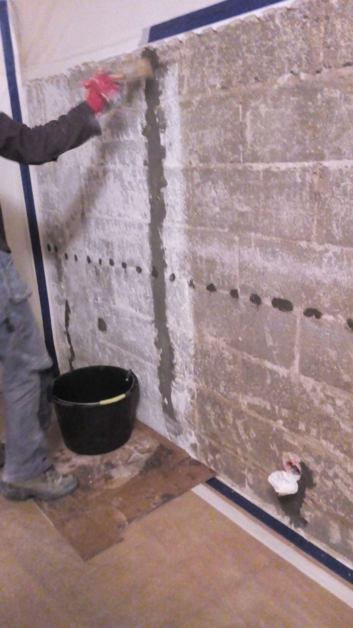 Mężczyzna w czerwonych rękawicach wypełnia zaprawą pionową szczelinę w ścianie z pustaków, widoczne nawiercone otwory, czarne wiadro i zabezpieczona folią podłoga.