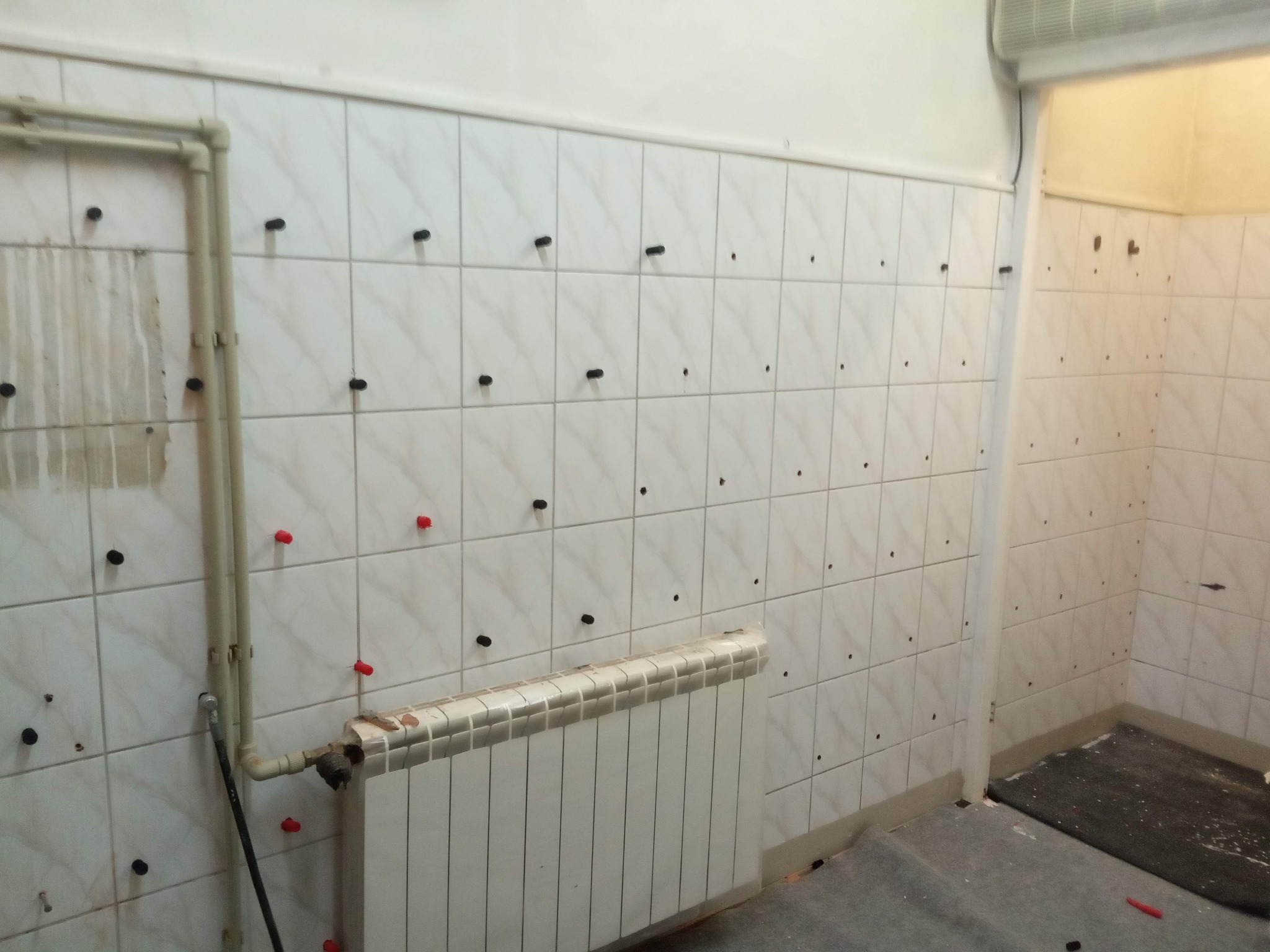 Świeżo położone, jasne płytki ścienne z czarnymi i czerwonymi krzyżykami dystansowymi, grzejnik i rury w trakcie prac wykończeniowych.