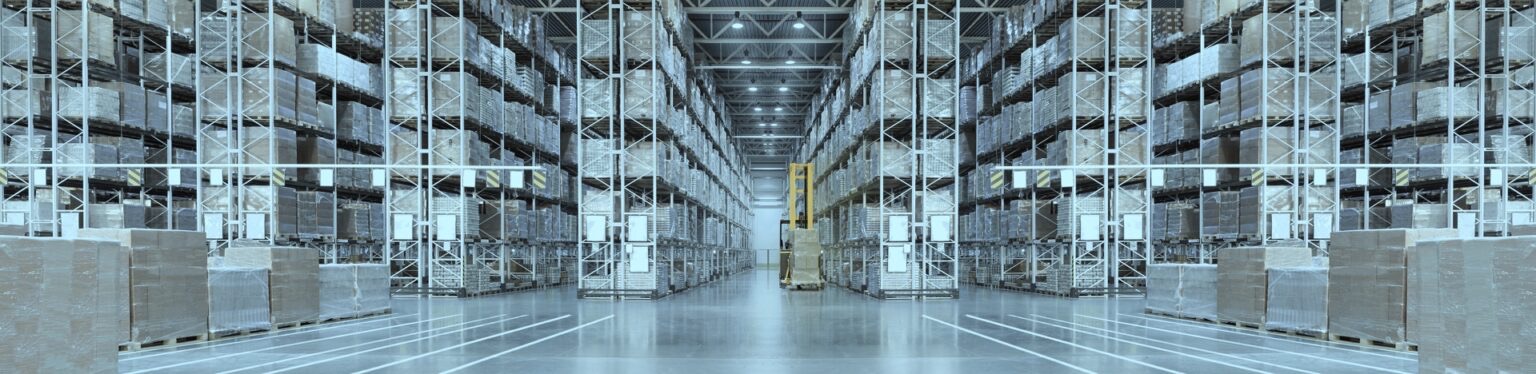 Wnętrze magazynu wysokiego składowania z regałami wypełnionymi zapakowanymi kartonami, posadzka z wyznaczonymi liniami, w tle widoczny wózek widłowy.