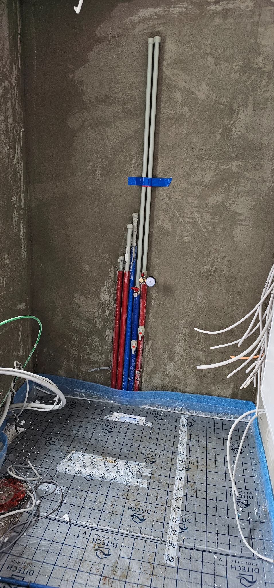 Wewnętrzne rury instalacji wodnej i kanalizacyjnej w trakcie montażu, częściowo w kolorze czerwonym i niebieskim, przymocowane taśmą do surowej ściany, widoczne także przewody elektryczne i mata...