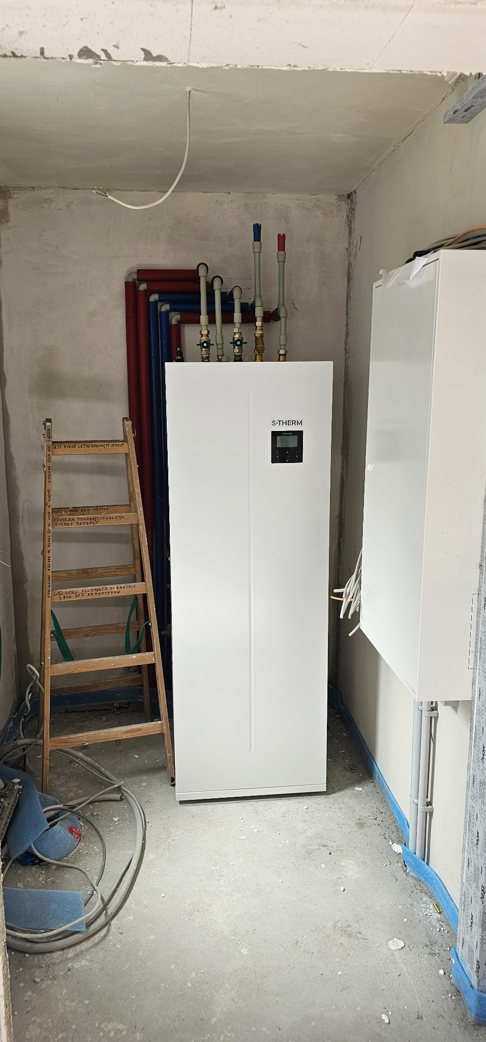 Biały, nowoczesny piec S-Therm z podłączonymi rurami wodnymi w surowym pomieszczeniu w trakcie instalacji. Obok pieca stoi drewniana drabina z napisami, a na podłodze leżą zwinięte kable.