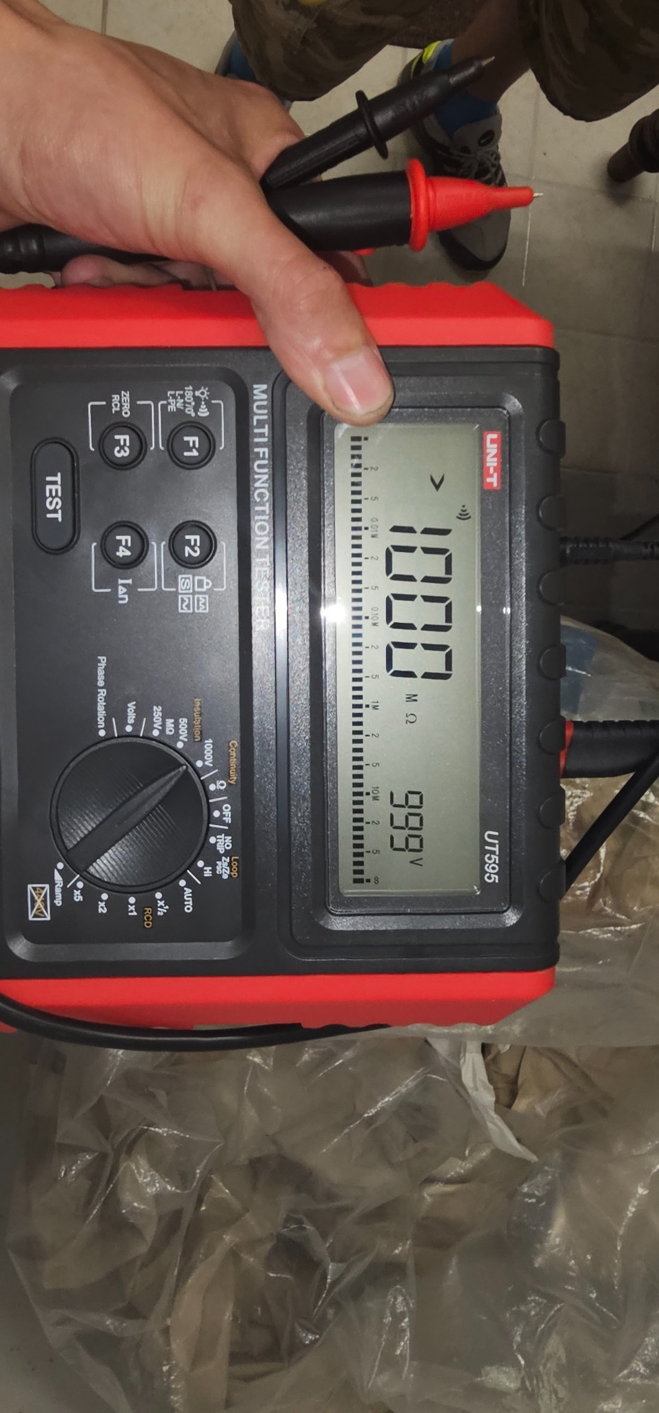 Miernik uniwersalny UT595 marki UNI-T, trzymany w dłoni, wyświetla wynik 1000 MΩ i 999 V, widoczne przewody pomiarowe z czarną i czerwoną końcówką, tło stanowi podłoga pokryta folią.